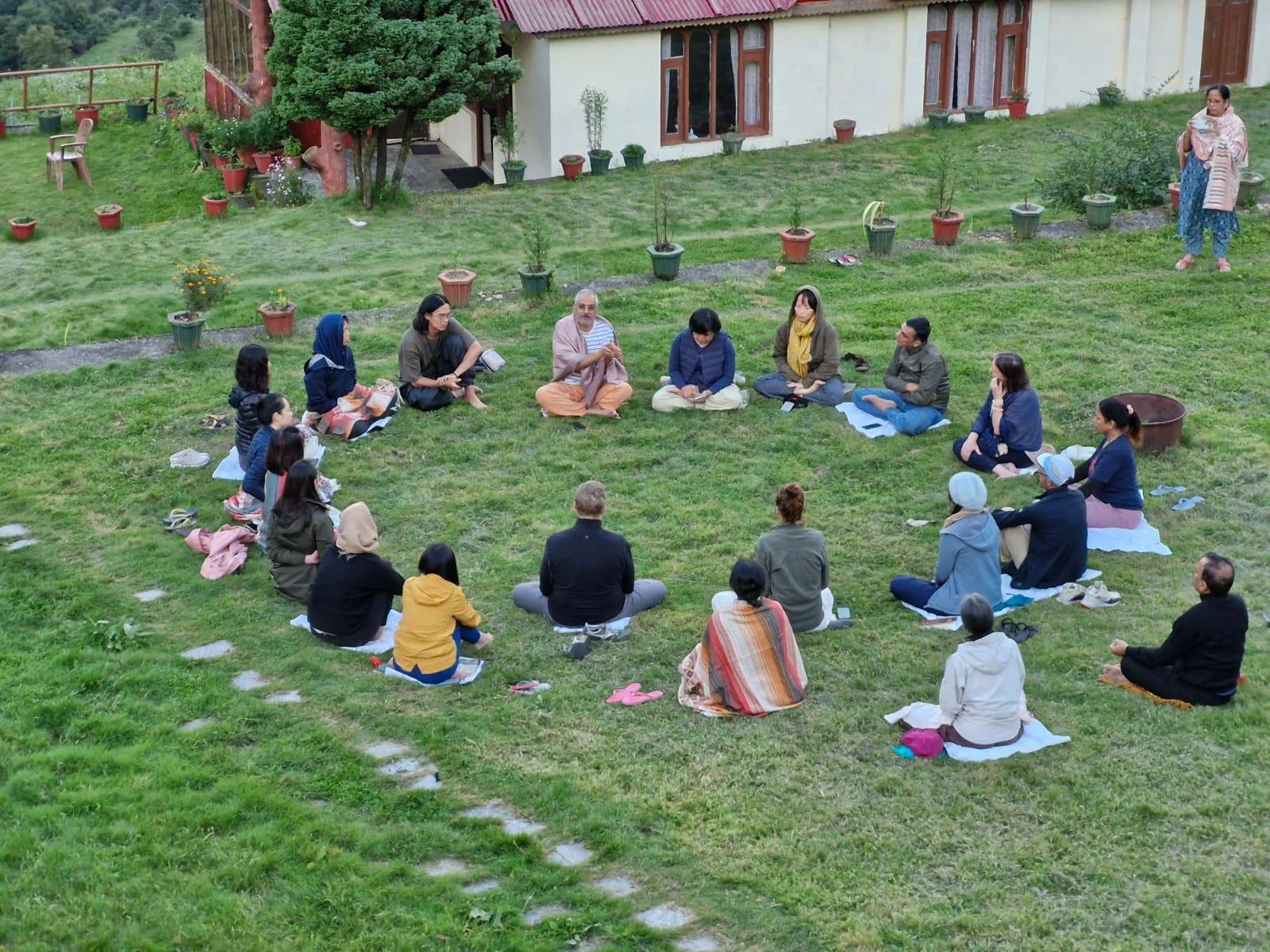 Gruppe von Menschen, die im Kreis auf einer Wiese sitzen, vermutlich bei einem Yoga- oder Meditationskurs, vor einem Haus mit Pflanzen und Bäumen.