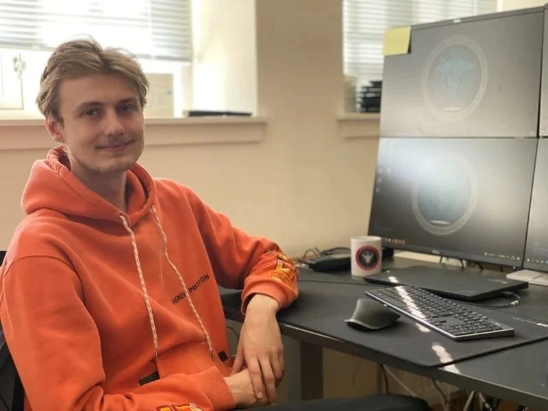 Ung mand sidder ved et skrivebord med tre computerskærme, et tastatur og en mus, iført en orange hoodie.