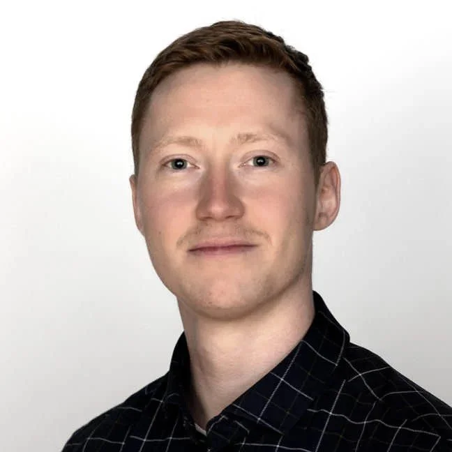 Portræt af en ung mand med lyst kort hår, let skæg, iført en sort skjorte med kvadratiske mønstre, mod en lysegrå baggrund.