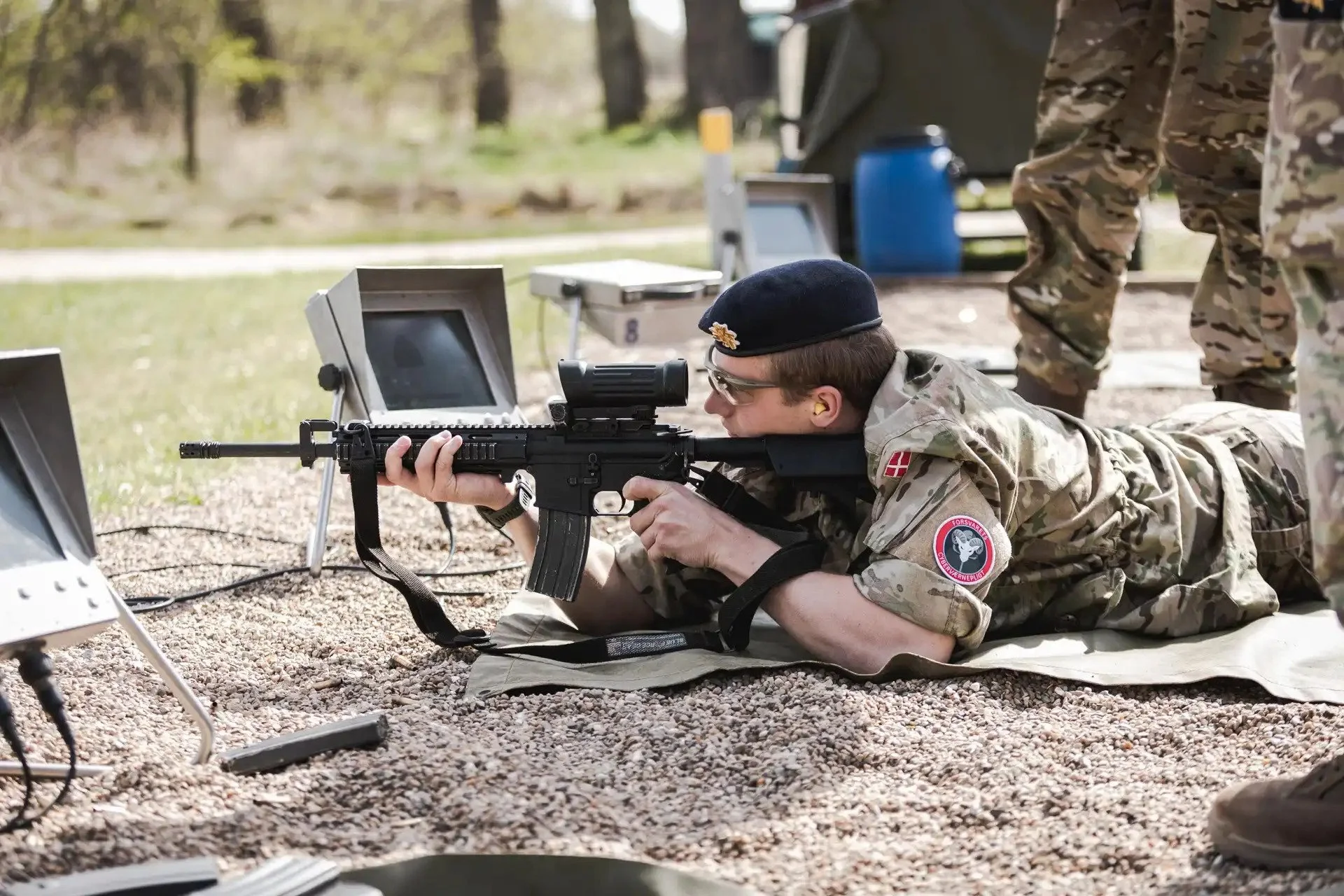 Soldat i camouflageuniform ligger på maven og skyder med et gevær under en skydeøvelse udenfor.