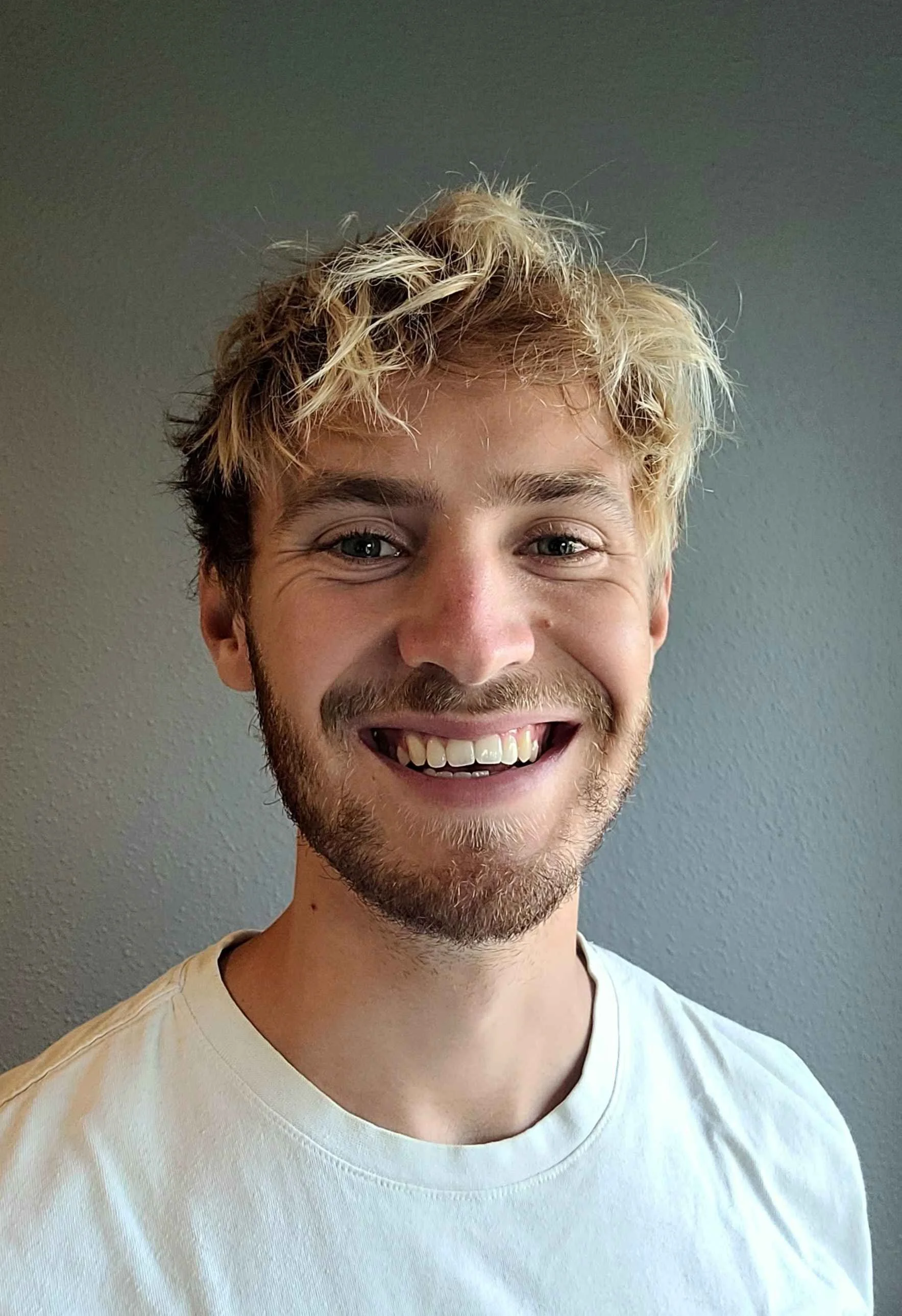 Ung mand med lyst, krøllet hår og skæg, smilende mod kameraet, iført hvid tshirt, stående foran en grå væg.