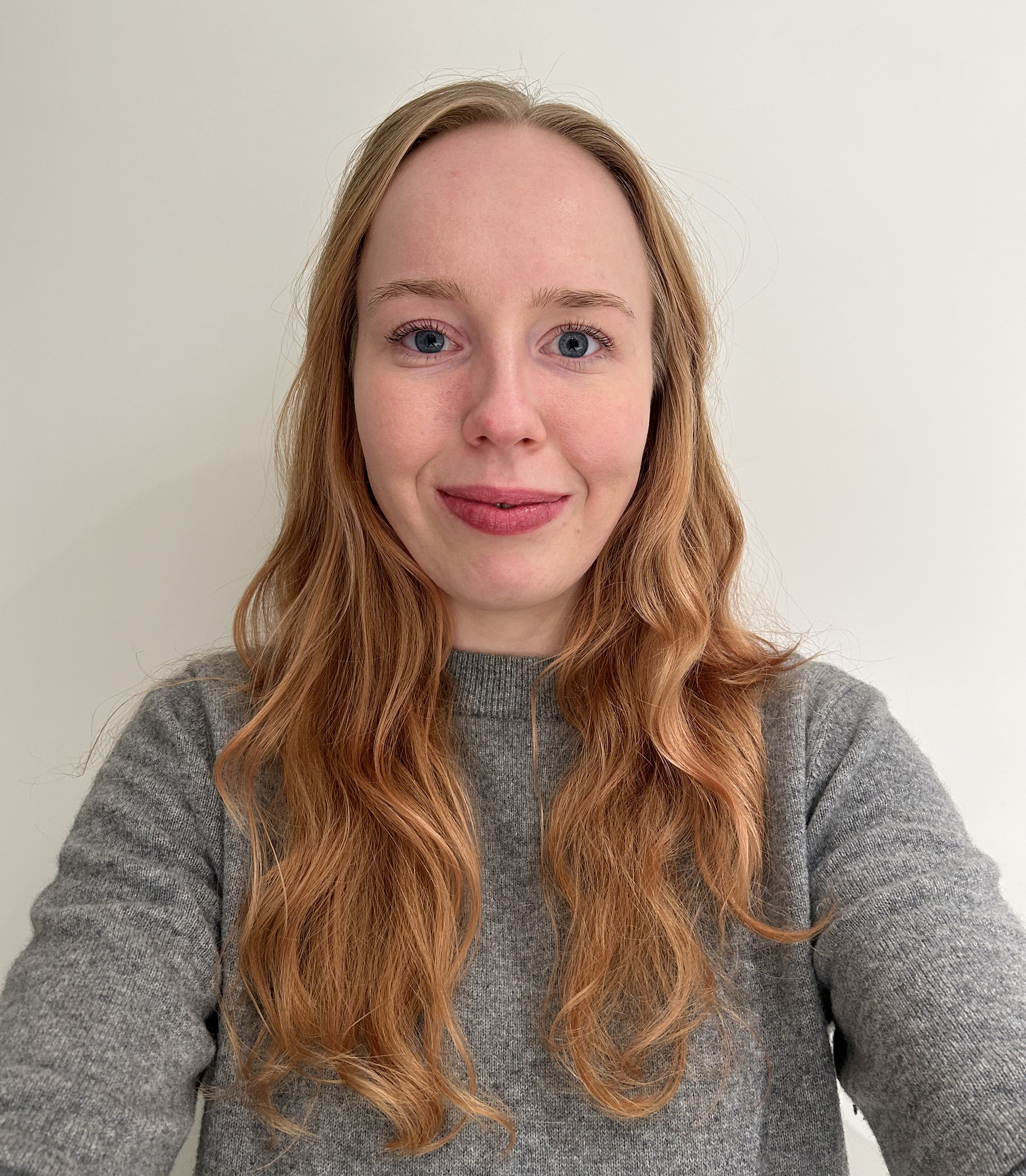 Et close-up foto af en kvinde med lang, krøllet, rødt hår og blå øjne, der smiler let, iført en grå sweater, mod en lysegrå væg.