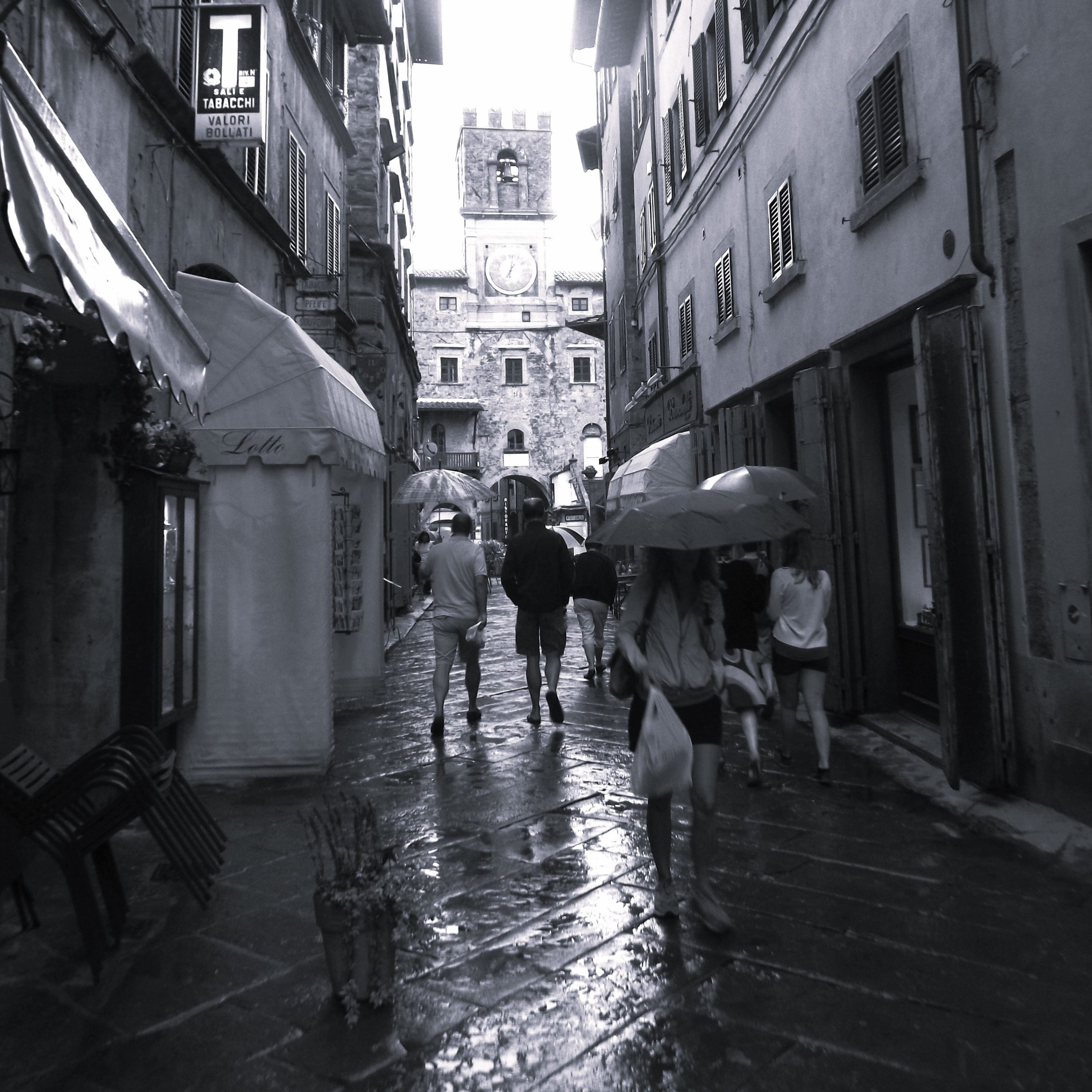 People walking with umbrellas on a wet, narrow street in an old European town with a clock tower in the background.