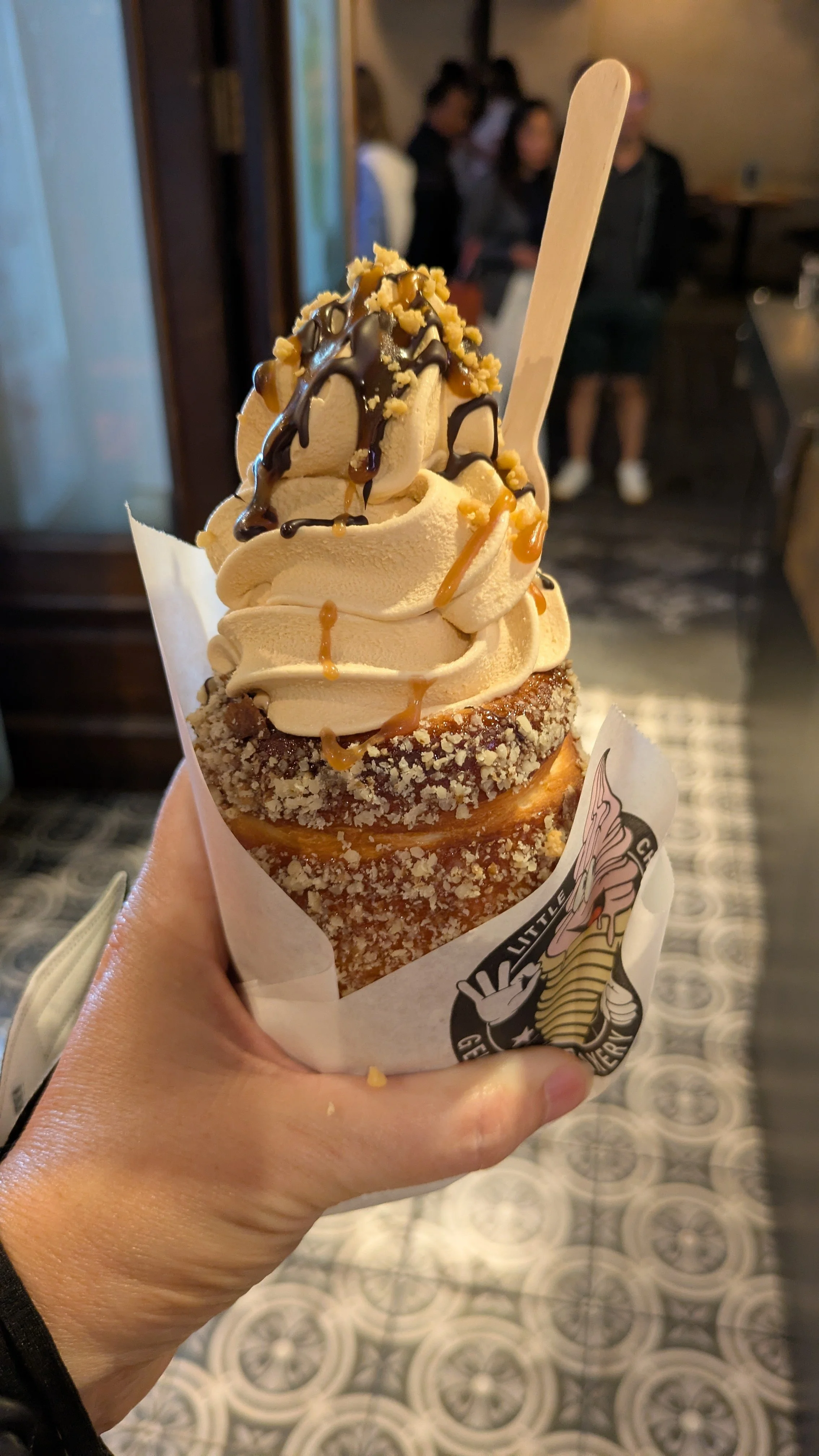 A hand holds a soft-serve ice cream with chocolate syrup and nuts, served in a pastry that looks like a rolled croissant.