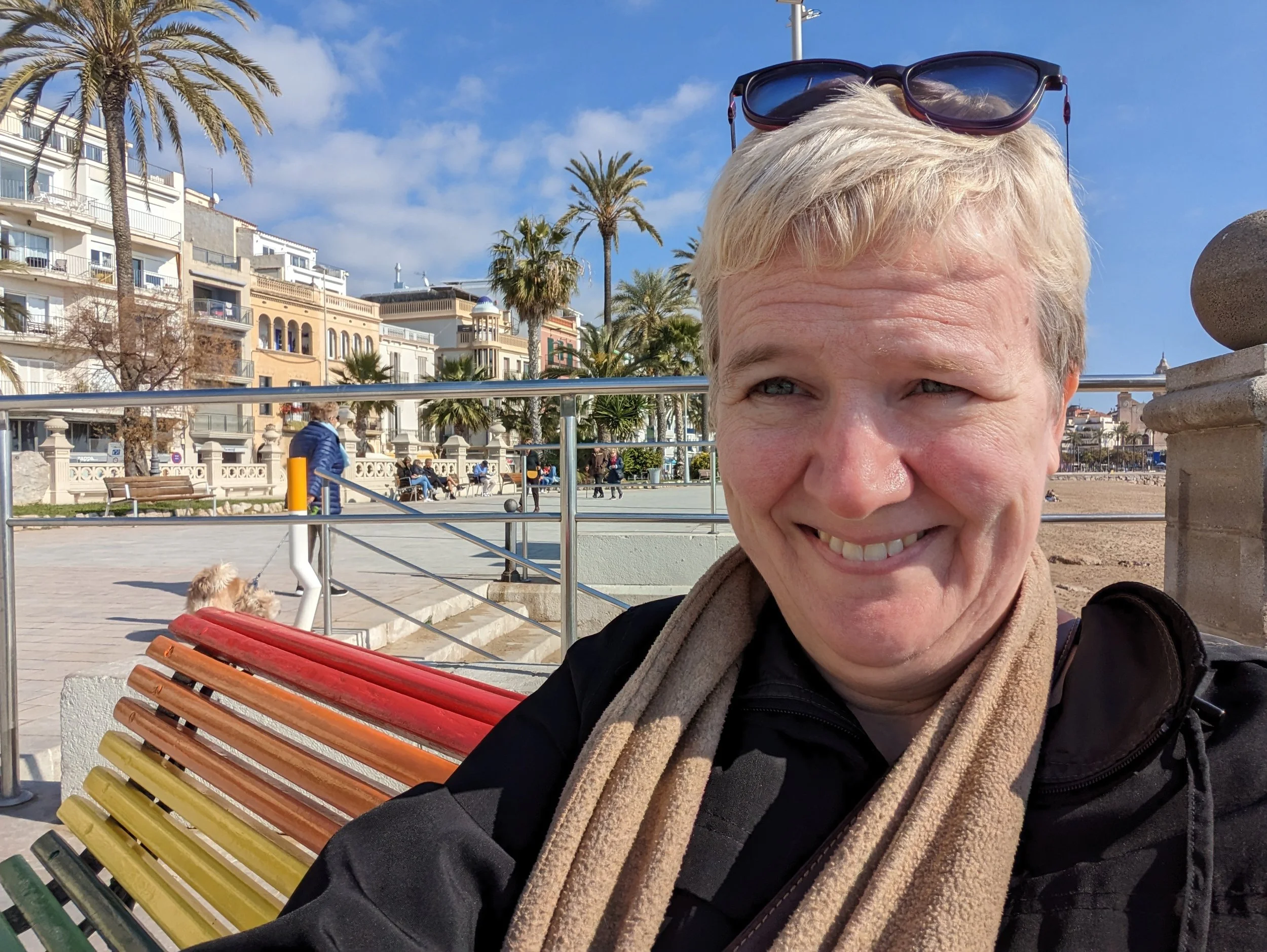 Blonde woman sitting on a rainbow striped bench.  Palm trees and buildings in the backgroun.
