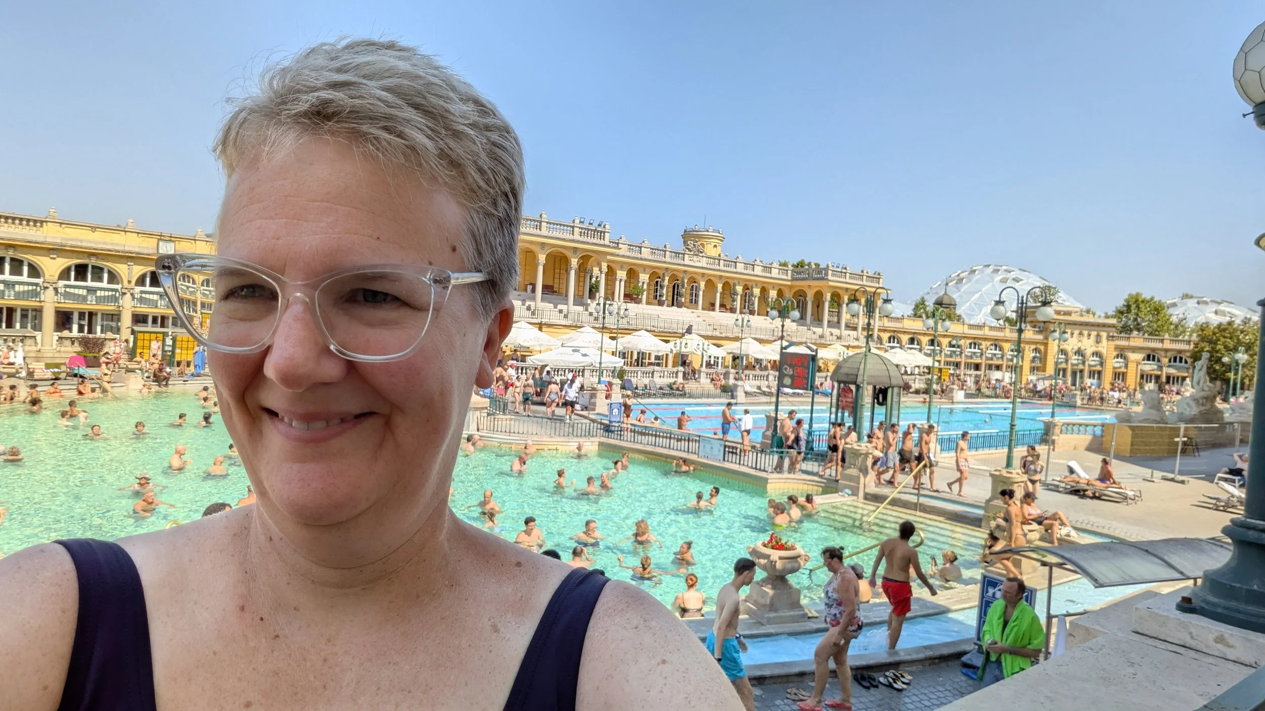 Short-haired blonde woman in bathing suit taking a selfie in front of a large swimming pool complex.