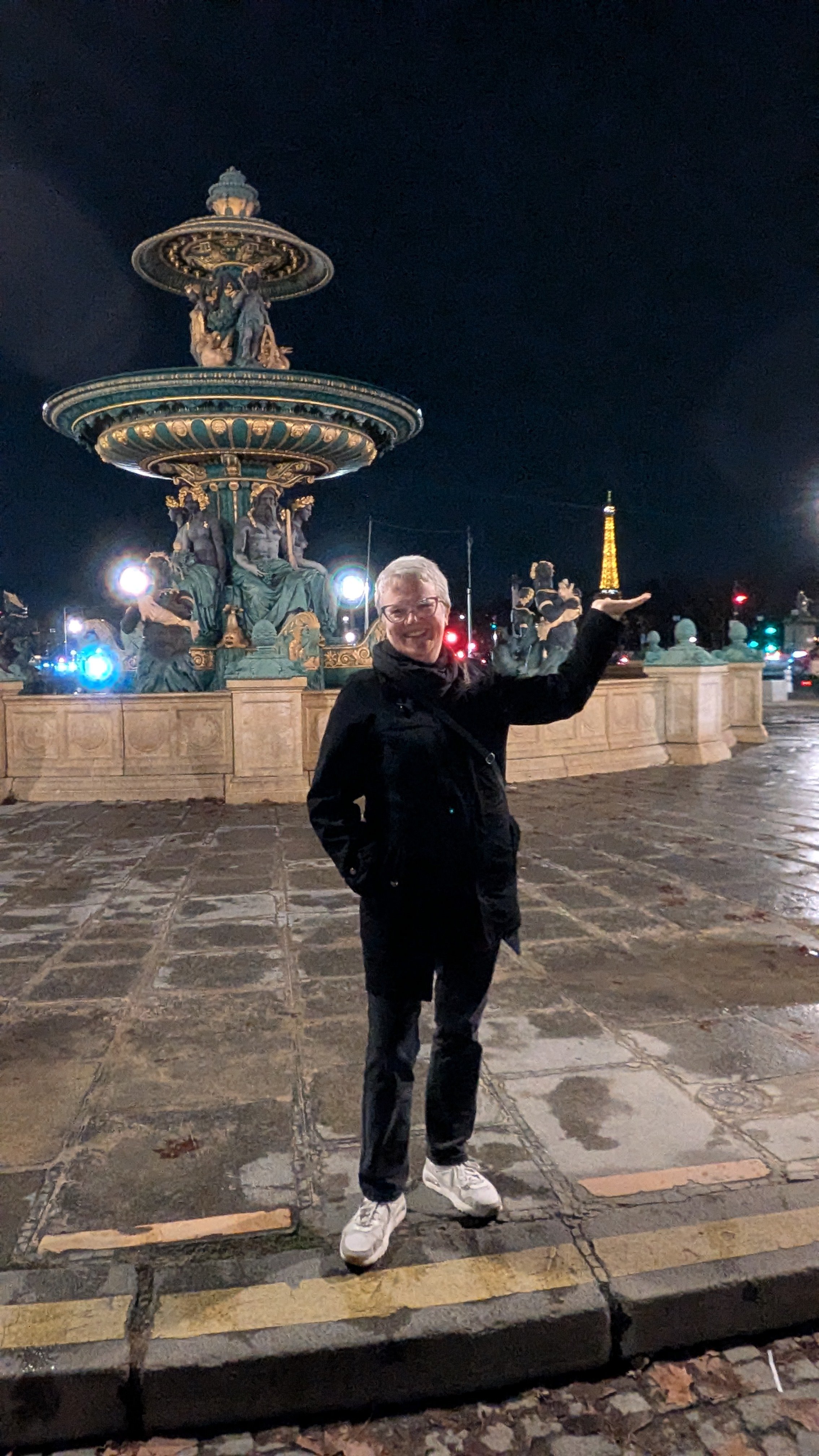 Woman in black coat and jeans stands in front of a copper and gold fountain, with the Eiffel Tower in the background.