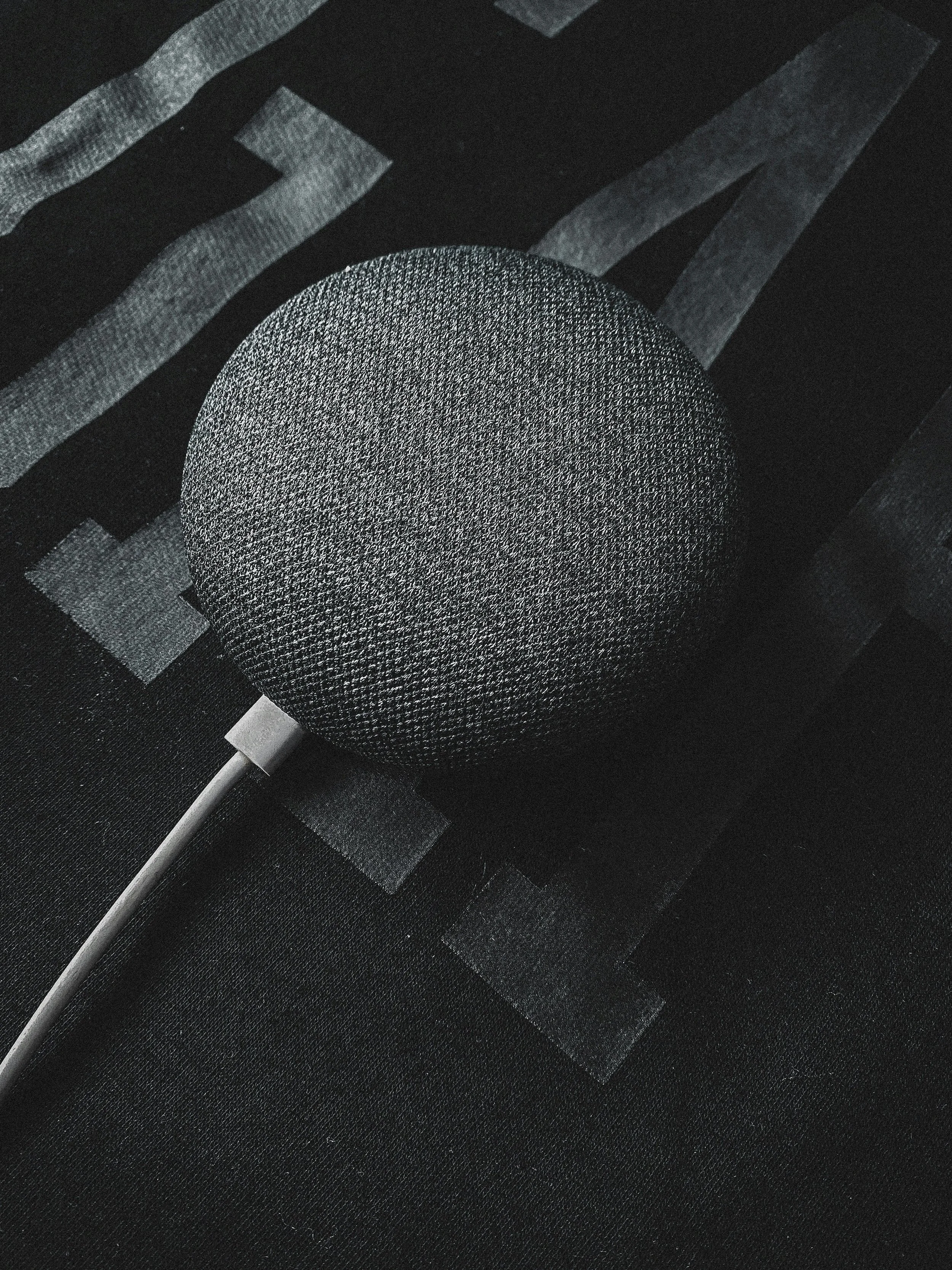 A black smart speaker device with a fabric cover and a cord, placed on a dark surface.