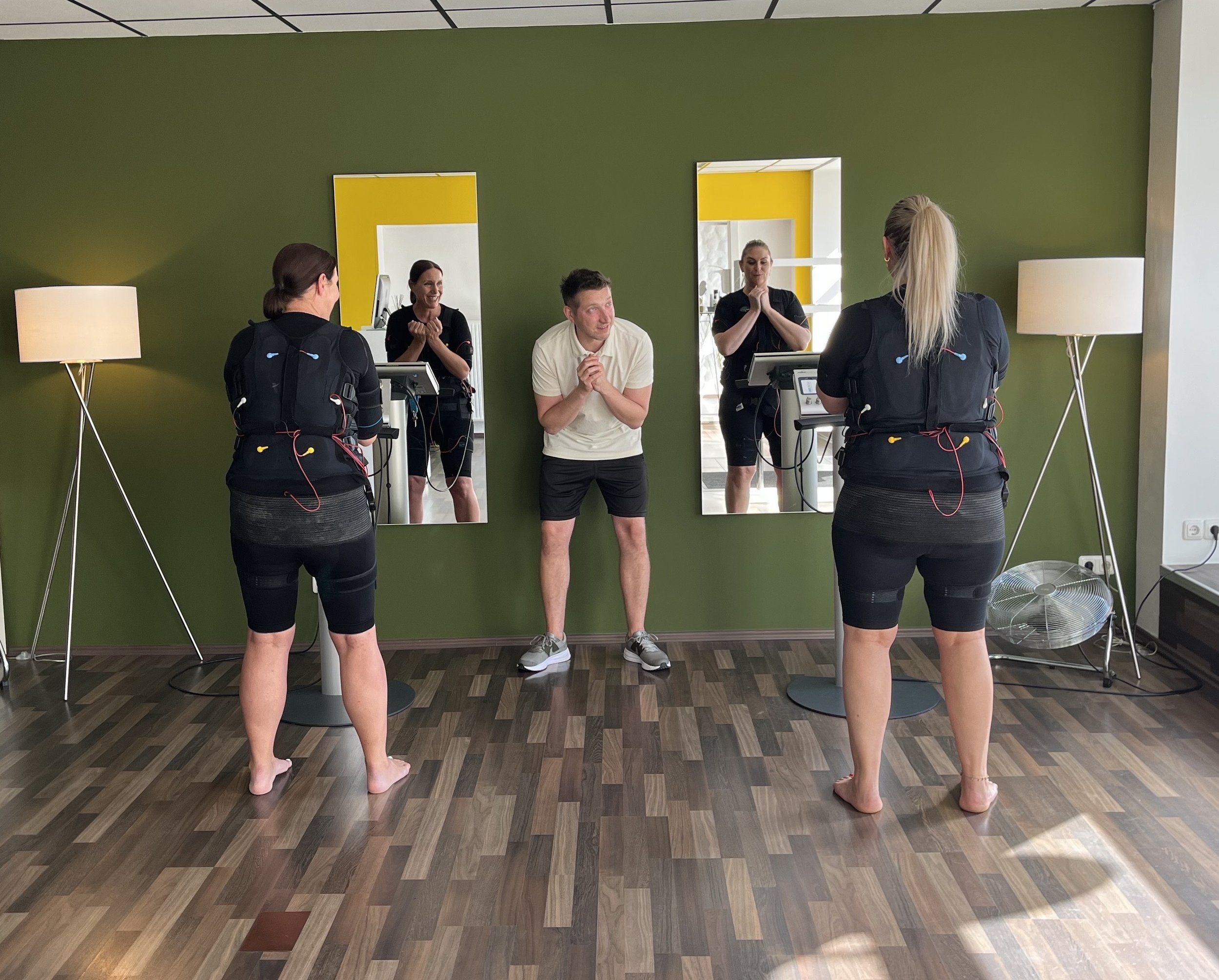 Zwei Frauen an elektrodenbasierten Kraftmessungen auf einem grünen Wand, ein Mann freut sich, während er in die Kameras lacht, die drei in einem Raum stehen.