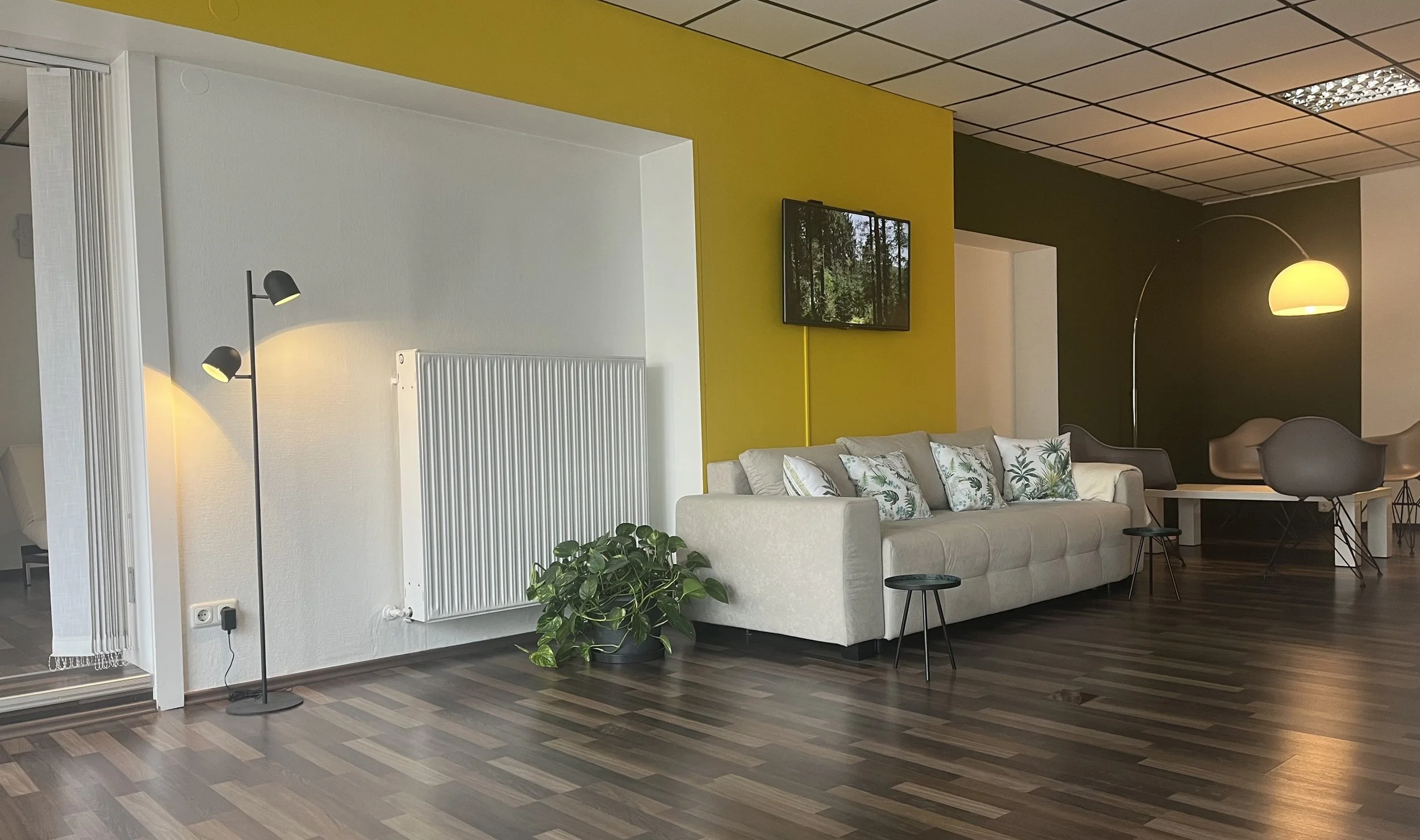 Modernes Wohnzimmer mit grauem Sofa, bunten Kissen, grünem Pflanzen, Standleuchte, Fernseher an gelber Wand und ovalen Sesselgruppen im Hintergrund, dunkler Wand und Lichter