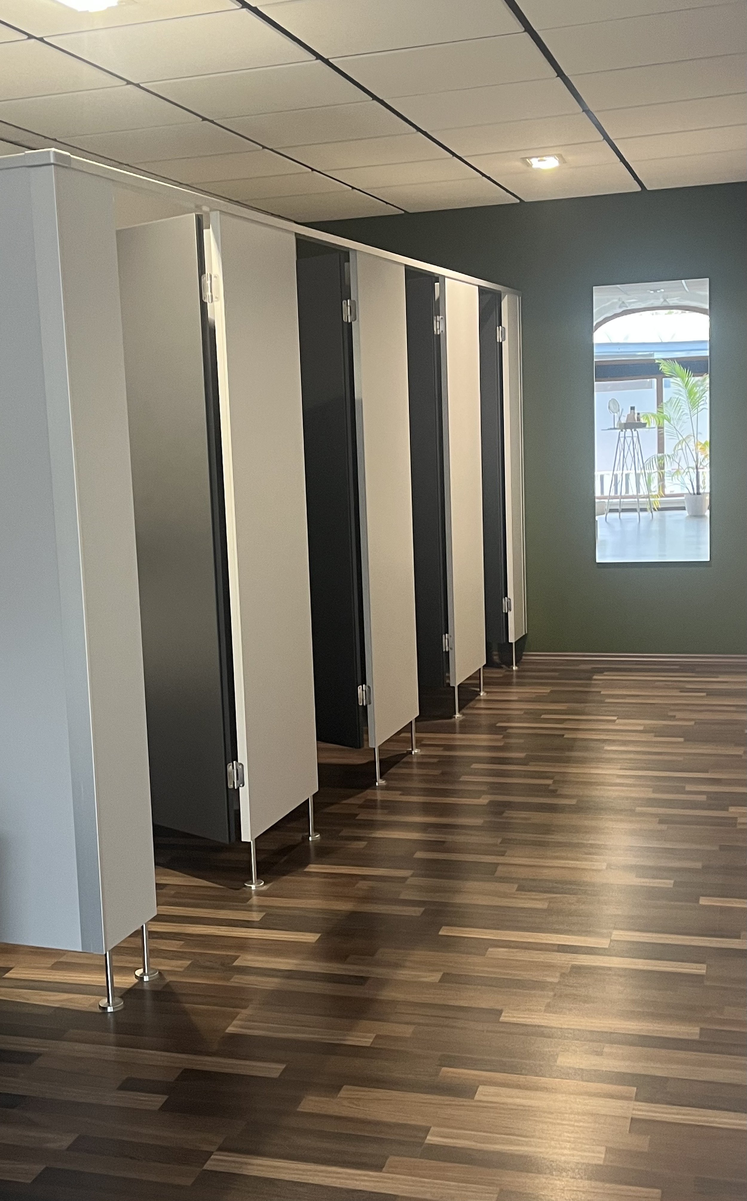 Umkleidekabine in einem Raum mit Holzboden und einem Fenster, durch das Pflanzen und ein Stehpult sichtbar sind.