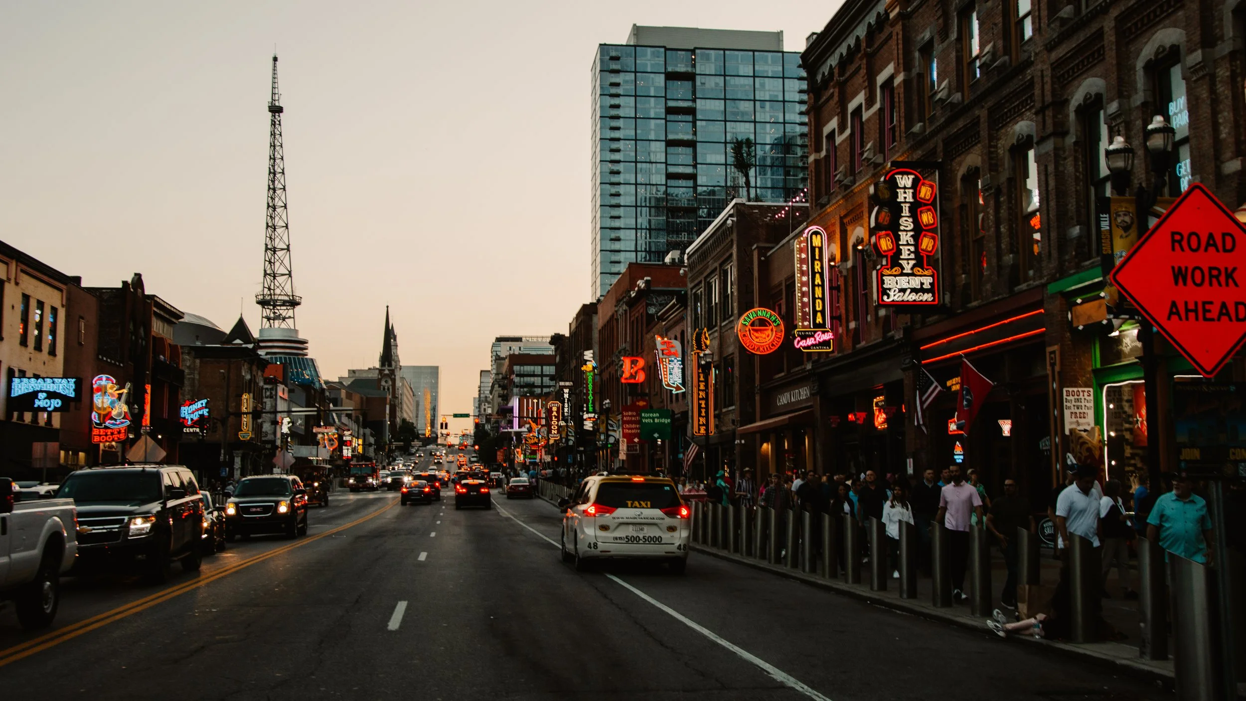 Busy street in an urban area with neon signs, bars, and restaurants, cars in traffic, and a prominent tower in the background; an orange "Road Work Ahead" sign is visible.