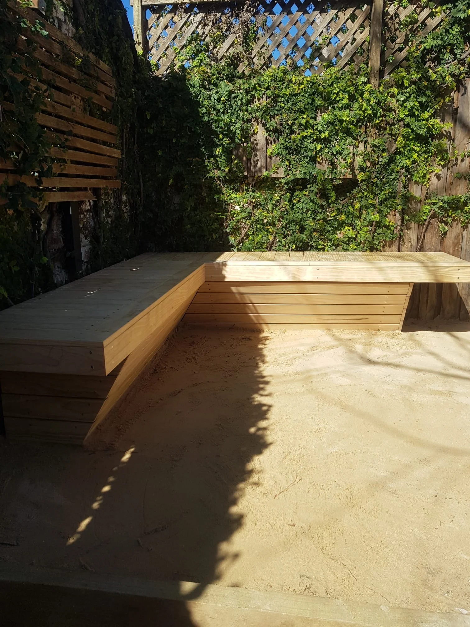 Wooden outdoor patio bench in a garden with sand ground and green leafy plants on the fence