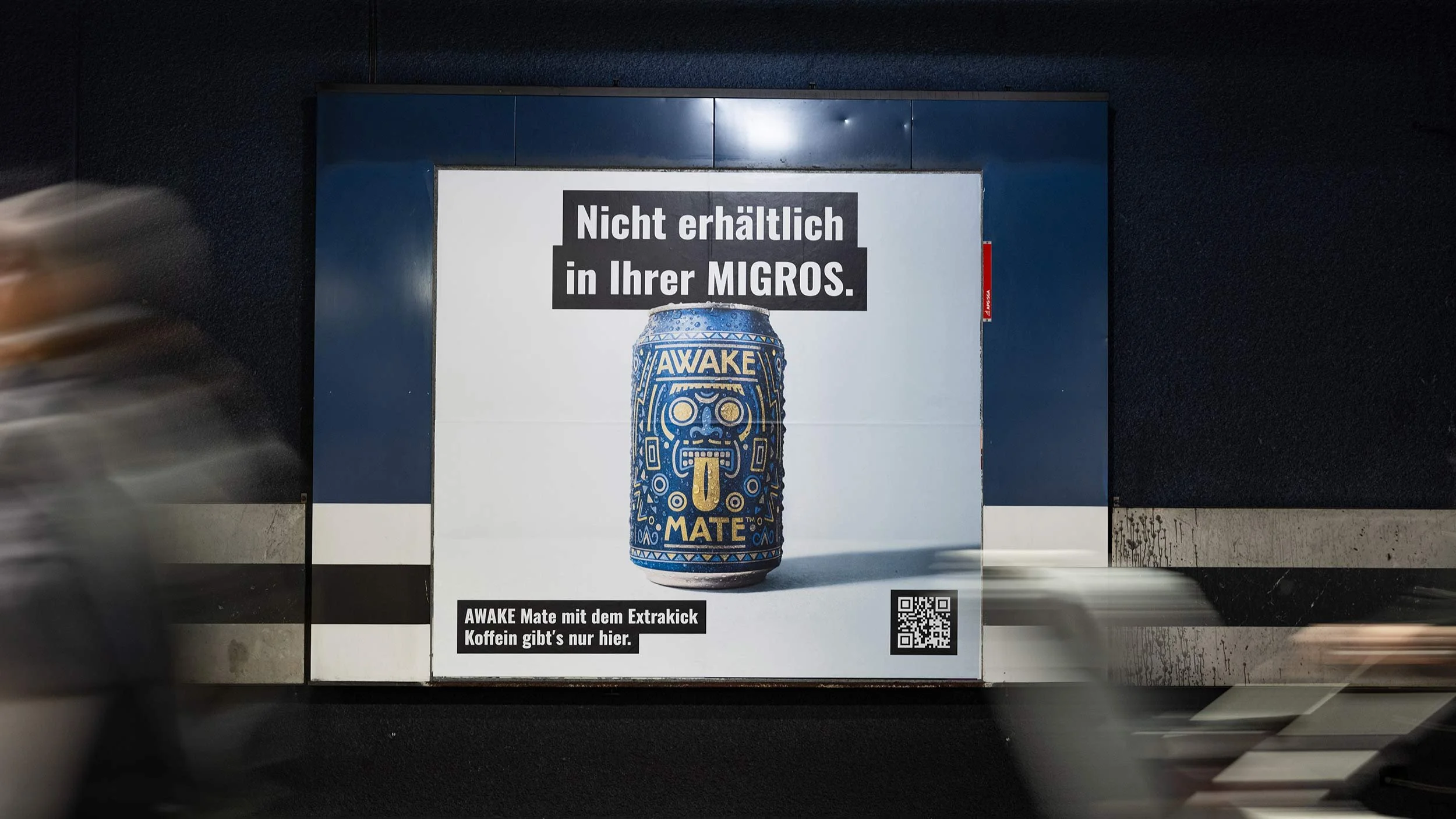 Werbung für WAKE Mate mit einem Dosen-Design, das eine an ethnischer Kunst inspirierte Illustration eines Gesichts zeigt. Der Text auf der Anzeige lautet: 'Nicht erhältich in Ihrer MIGROS.' und darunter: 'WAKE Mate mit dem Extrakick Koffein gibt's nur hier.' Ein QR-Code ist ebenfalls auf der Anzeige sichtbar.