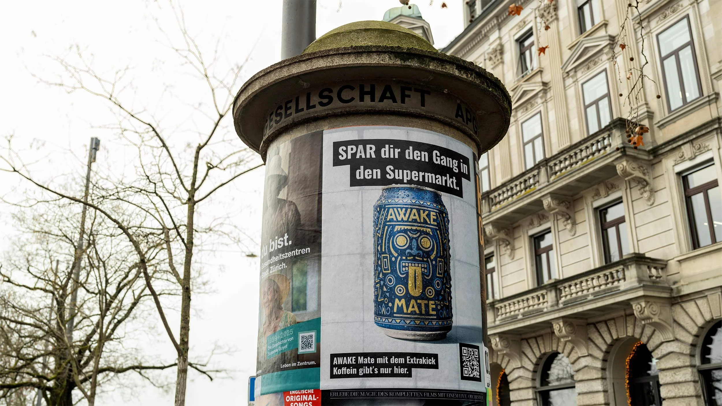 Werbung auf einem städtischen Werbecontainer, die einen Energydrink namens 'AWAKE' mit dem Slogan 'AWAKE Mate mit dem Extrakick Koffein gibt's nur hier' zeigt, im Hintergrund eine historische Gebäudefassade, trockene Baumzweige.
