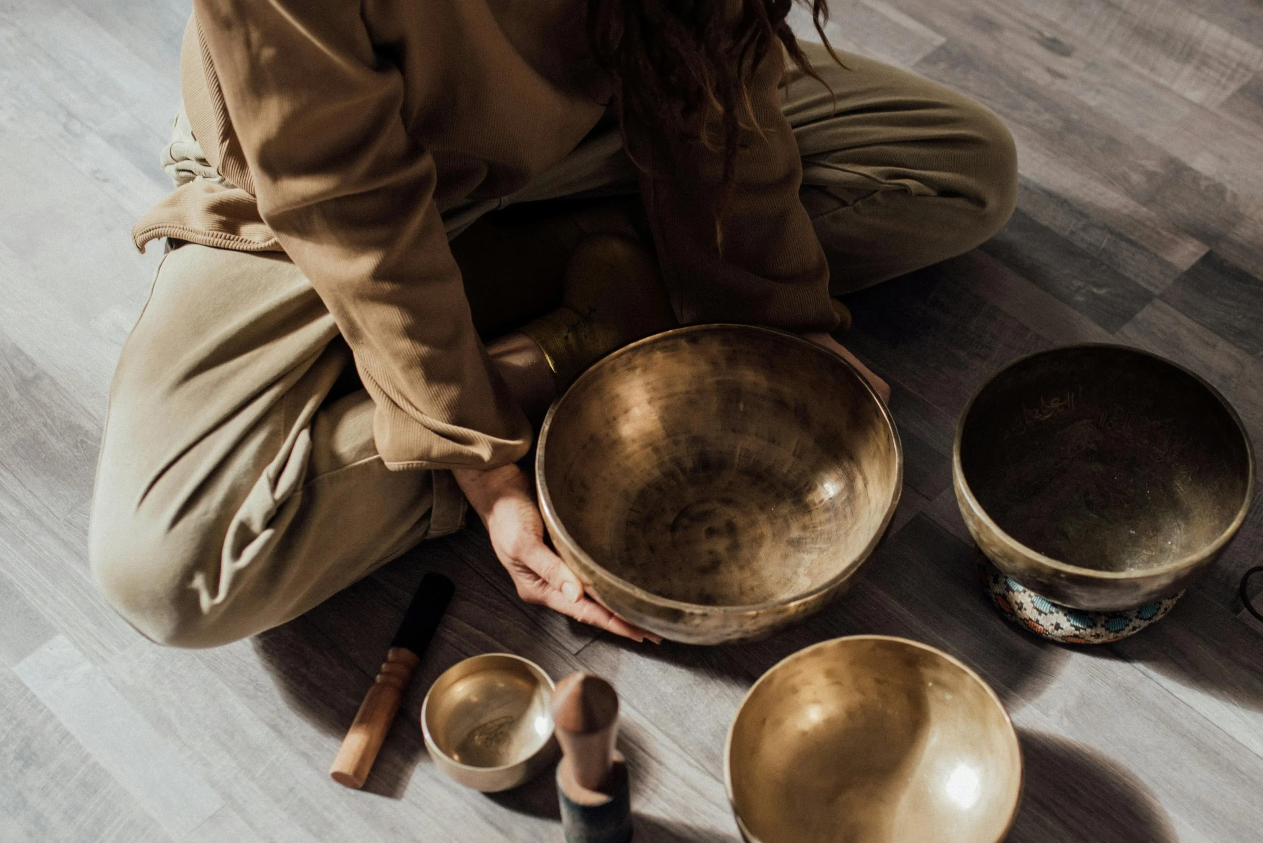 Person sitting on the floor holding a metallic singing bowl with other similar bowls around. The person is wearing beige clothing and brown shoes, with long hair.