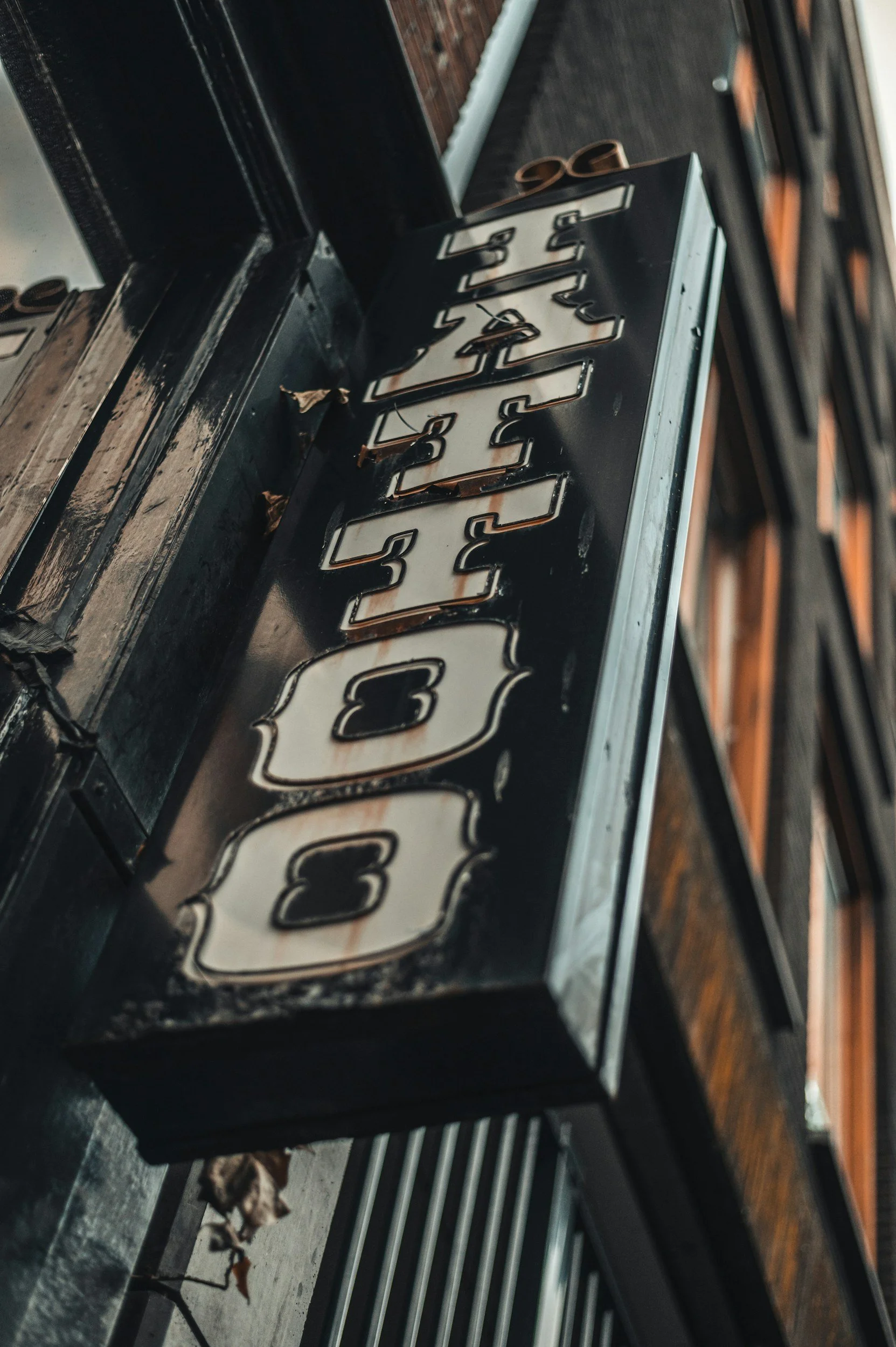 Close-up of an illuminated vintage neon sign that reads 'COFFEE', with parts of the sign showing rust and weathering, mounted on a dark wall, with dried leaves and debris scattered nearby.