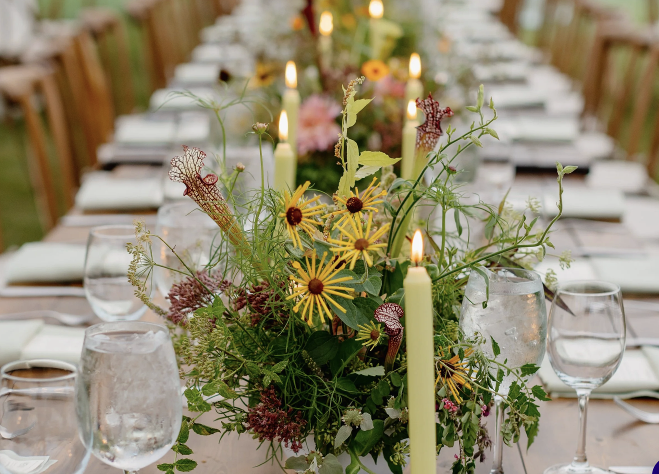 A long dining table set for a special event, decorated with a lush floral centerpiece, lit candles, glassware, and arranged with napkins and plates.