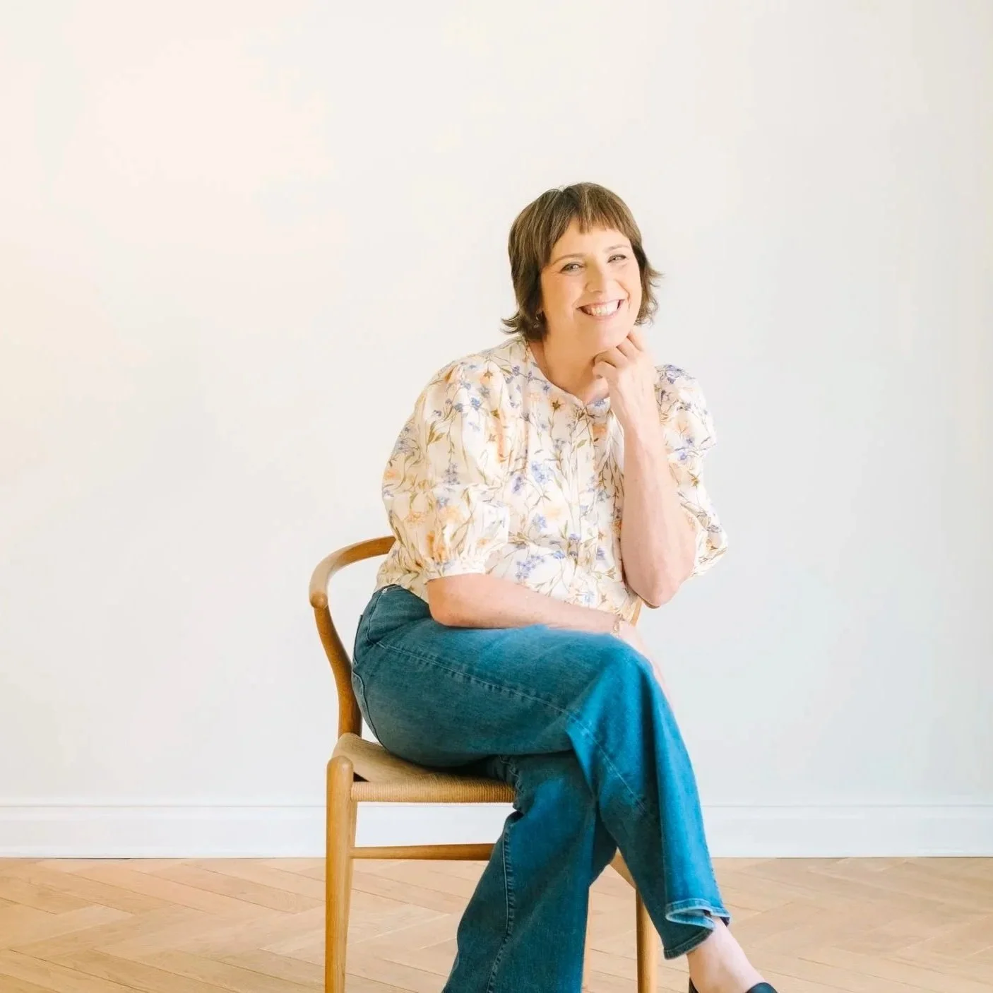 woman sitting on a chair with legs crossed wearing a floral blouse and blue jeans