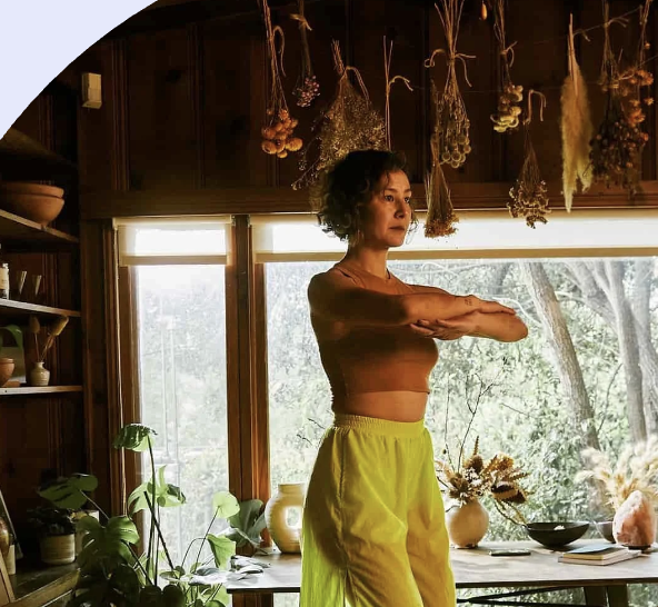 A woman standing indoors next to windows with a forest view, stretching her arms forward while looking to the side. The room has wooden walls and shelves, with hanging dried herbs and plants above.