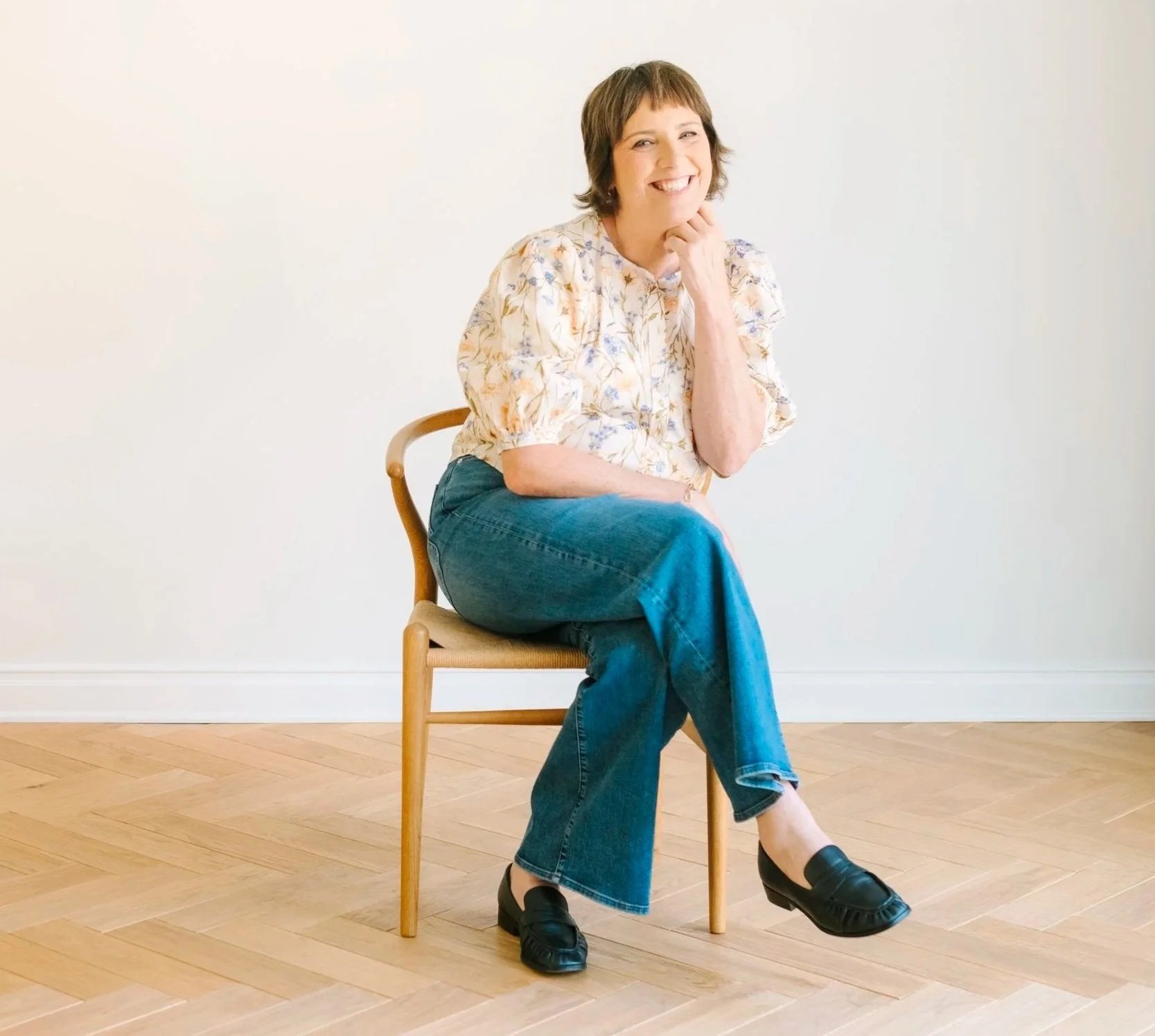 A woman with short brown hair sitting on a wooden chair with a beige seat in front of a plain white wall, smiling and resting her chin on her hand. She is wearing a floral blouse, blue jeans, and black slip-on shoes.