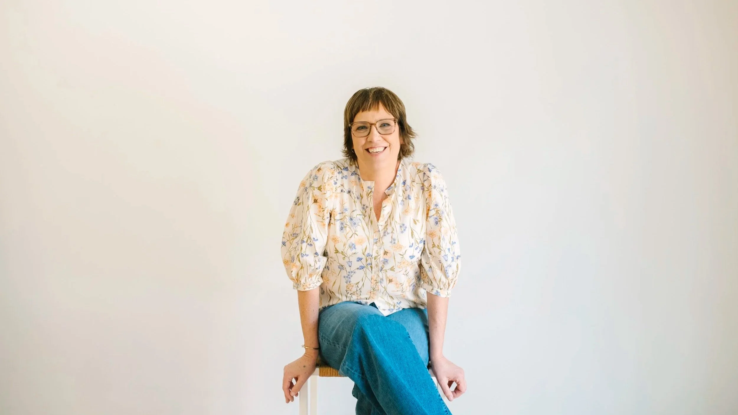 woman sitting on chair wearing brown glasses, floral shirt and blue jeans
