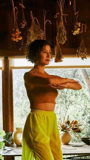 a woman standing arms folded with dried flowers hanging from the ceiling and a large window overlooking a garden