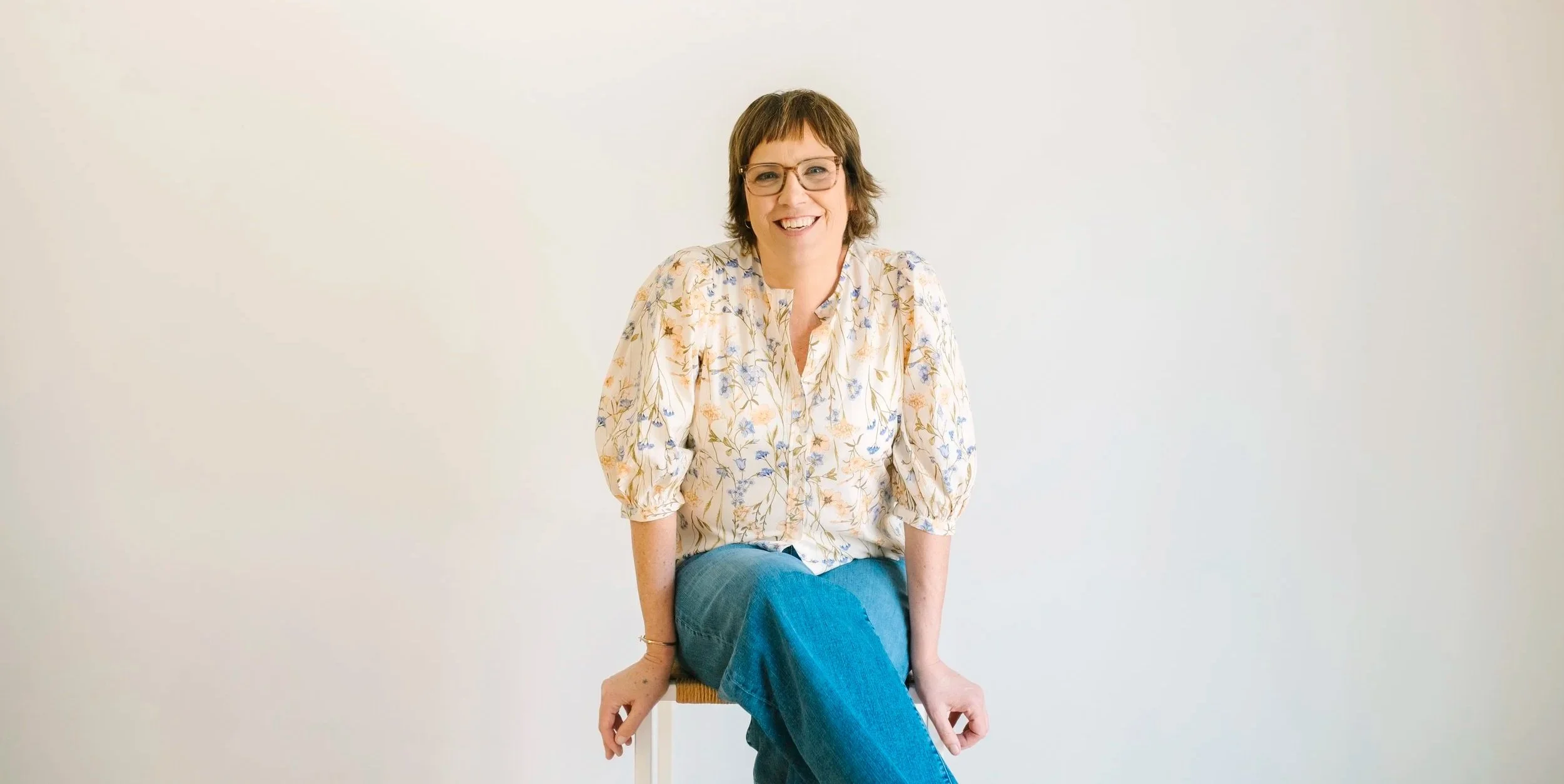 woman sitting on stool wearing blue jeans and floral shirt with brown hair and glasses