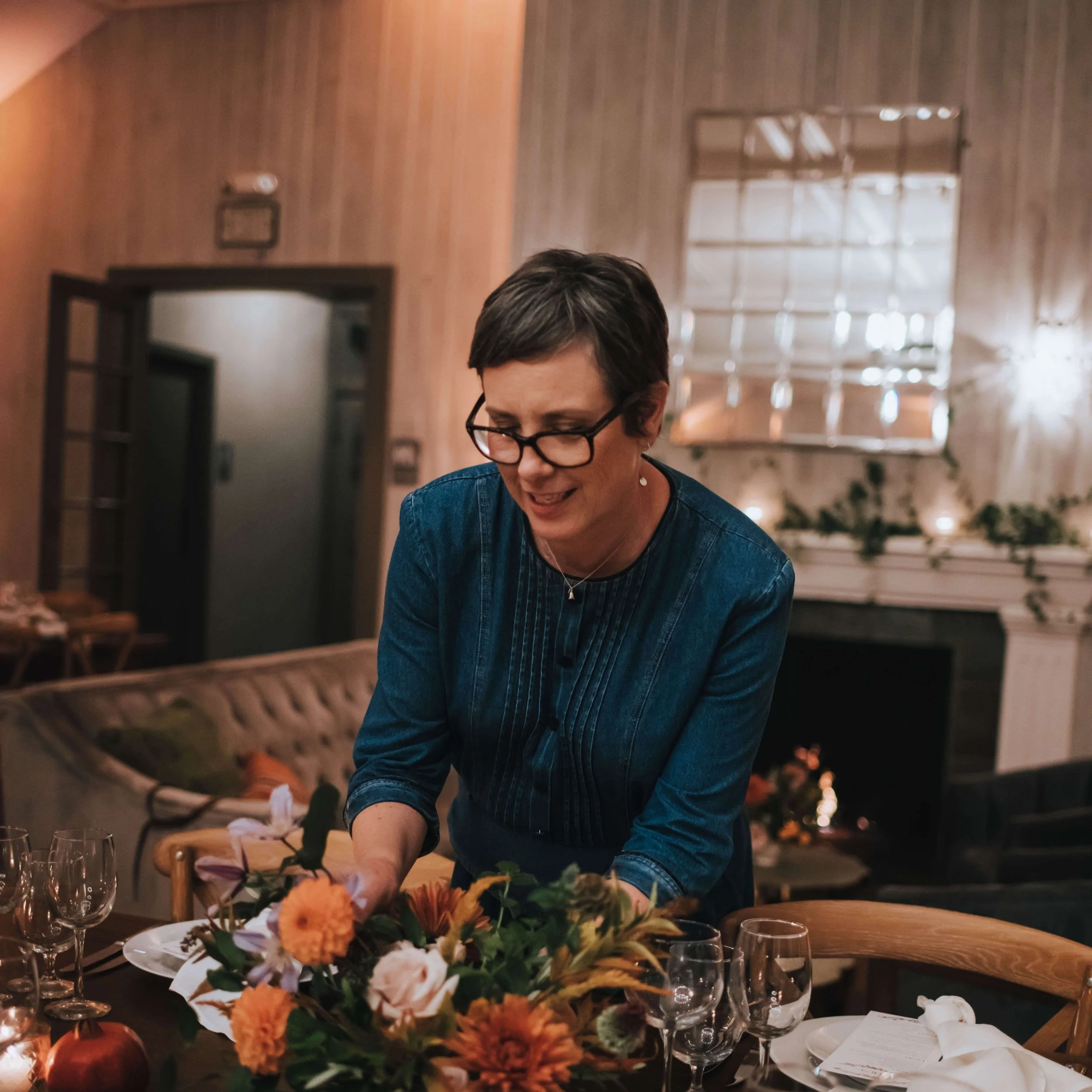woman wearing glasses and blue denim dress arranging flowers in a room with white walls and sofa
