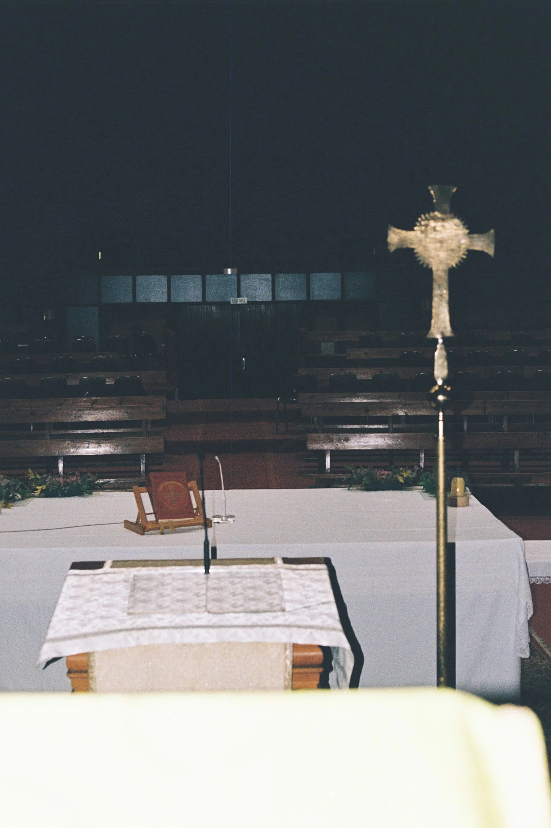 Escenario de misa en una iglesia, con altar cubierto por un mantón blanco, un libro y un candelabro. En el fondo, bancos y ventanales oscuros, y en el lateral, un crucifijo de piedra.