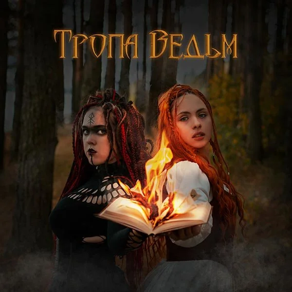 Two women with dressed in gothic style, one with black and red dreadlocks and the other with red hair, holding an open flaming book in a dark forest.