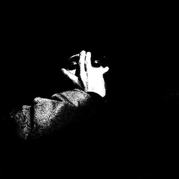 Person holding their hand up to their face in front of their eye, with the eye visible through fingers, in a black-and-white, high-contrast photo.
