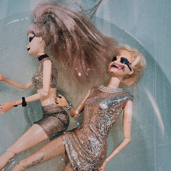 Two fashion dolls lying in a circle in water, with long hair and wearing shimmering outfits.