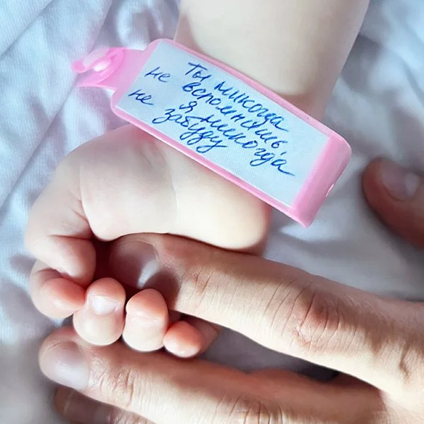 A person's hand with a pink wristband that has handwritten text in Russian, resting on a white fabric surface.