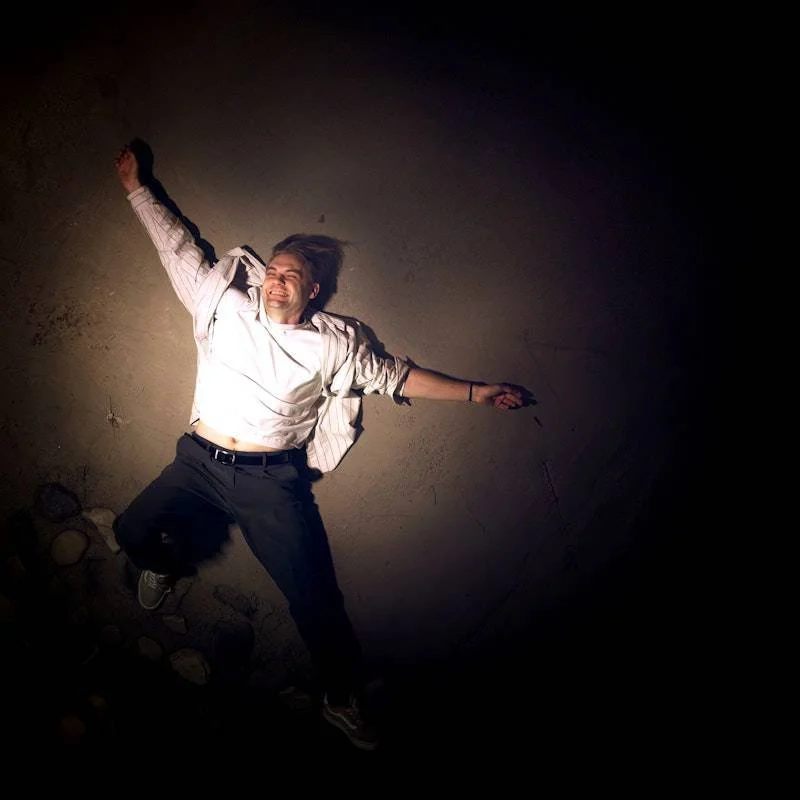Person lying on the ground smiling, illuminated by a camera flash, with arms outstretched, on a concrete floor.