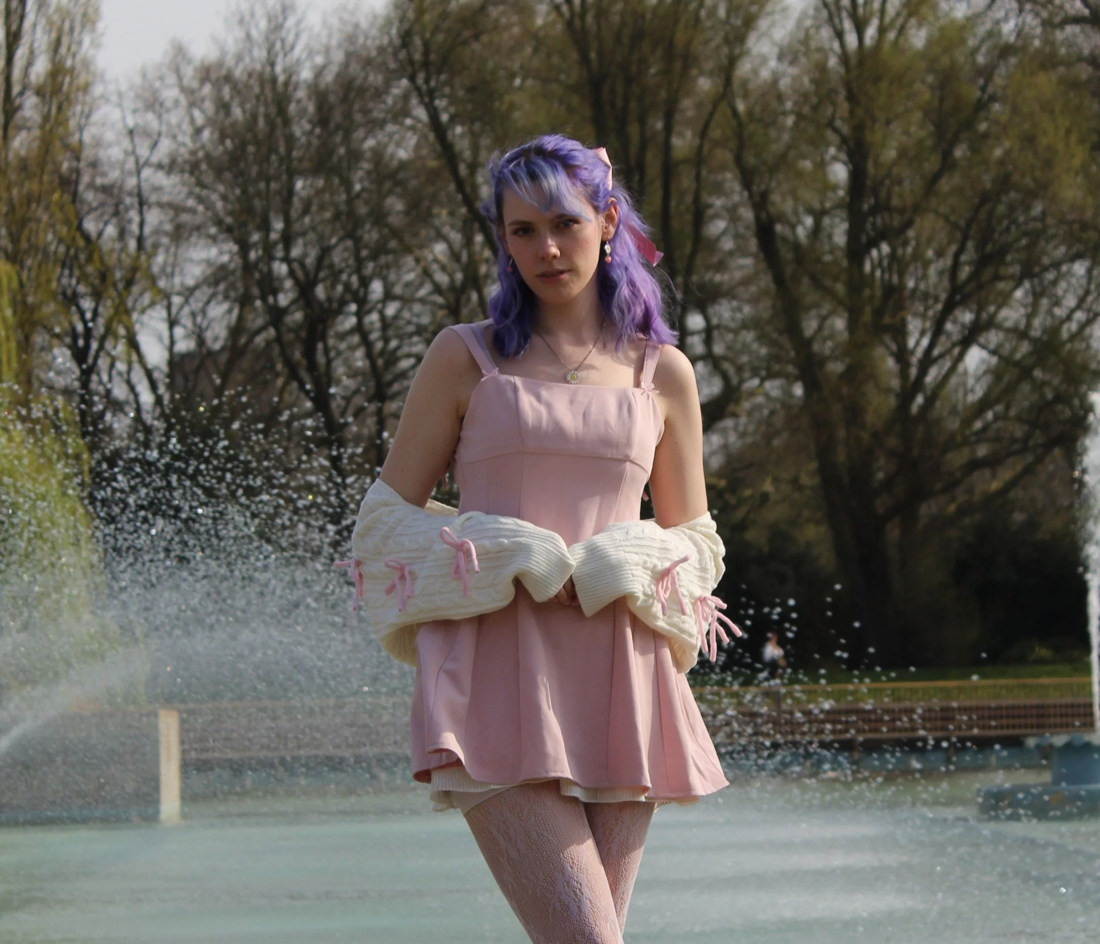 A woman with purple hair in a pink dress standing outdoors near water, holding multiple beige sweaters with pink ribbons, with trees in the background.