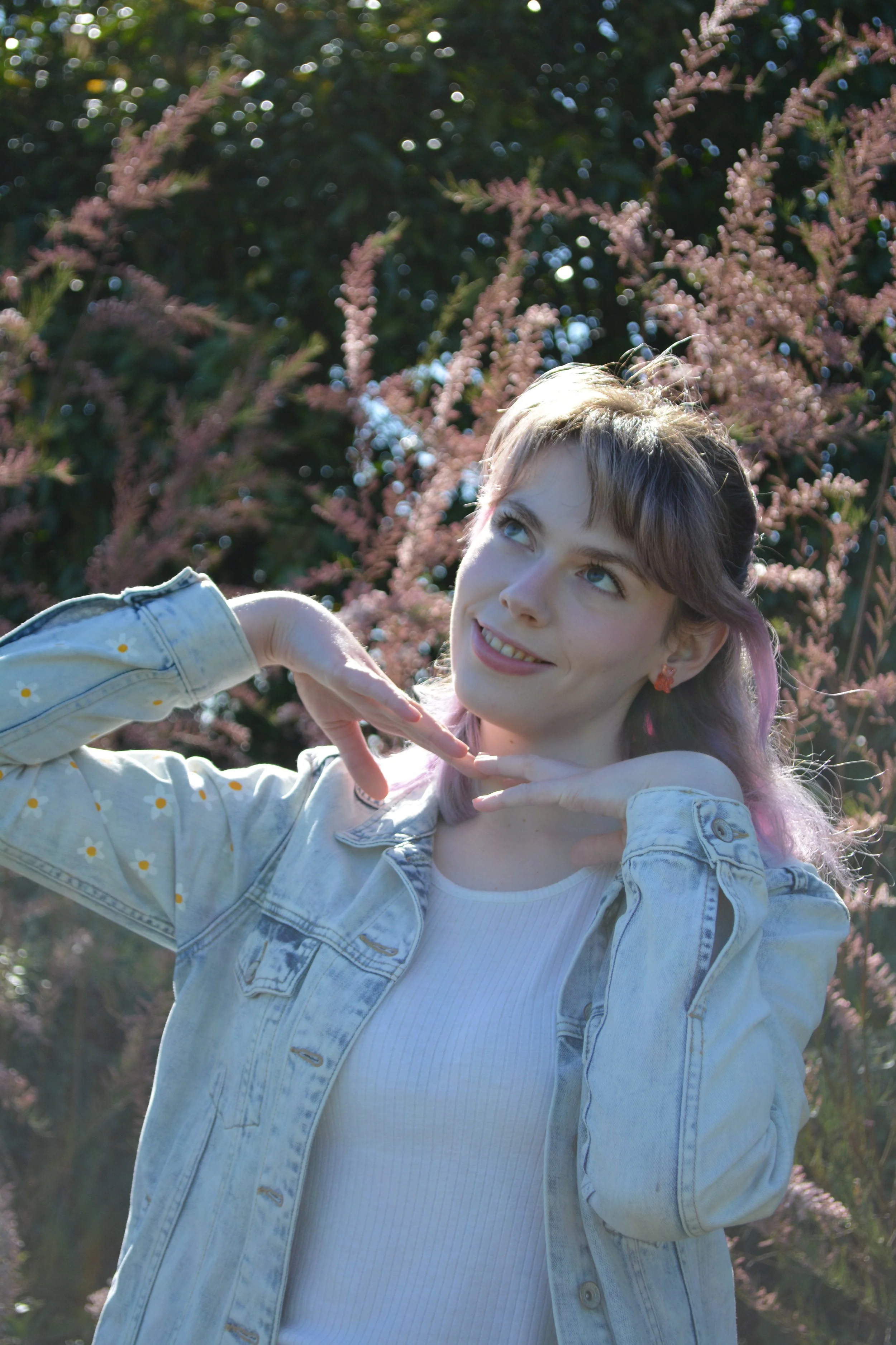 A young woman with purple hair and blue eyes smiling and making a heart shape with her hands in front of pink flowering bushes in sunlight.