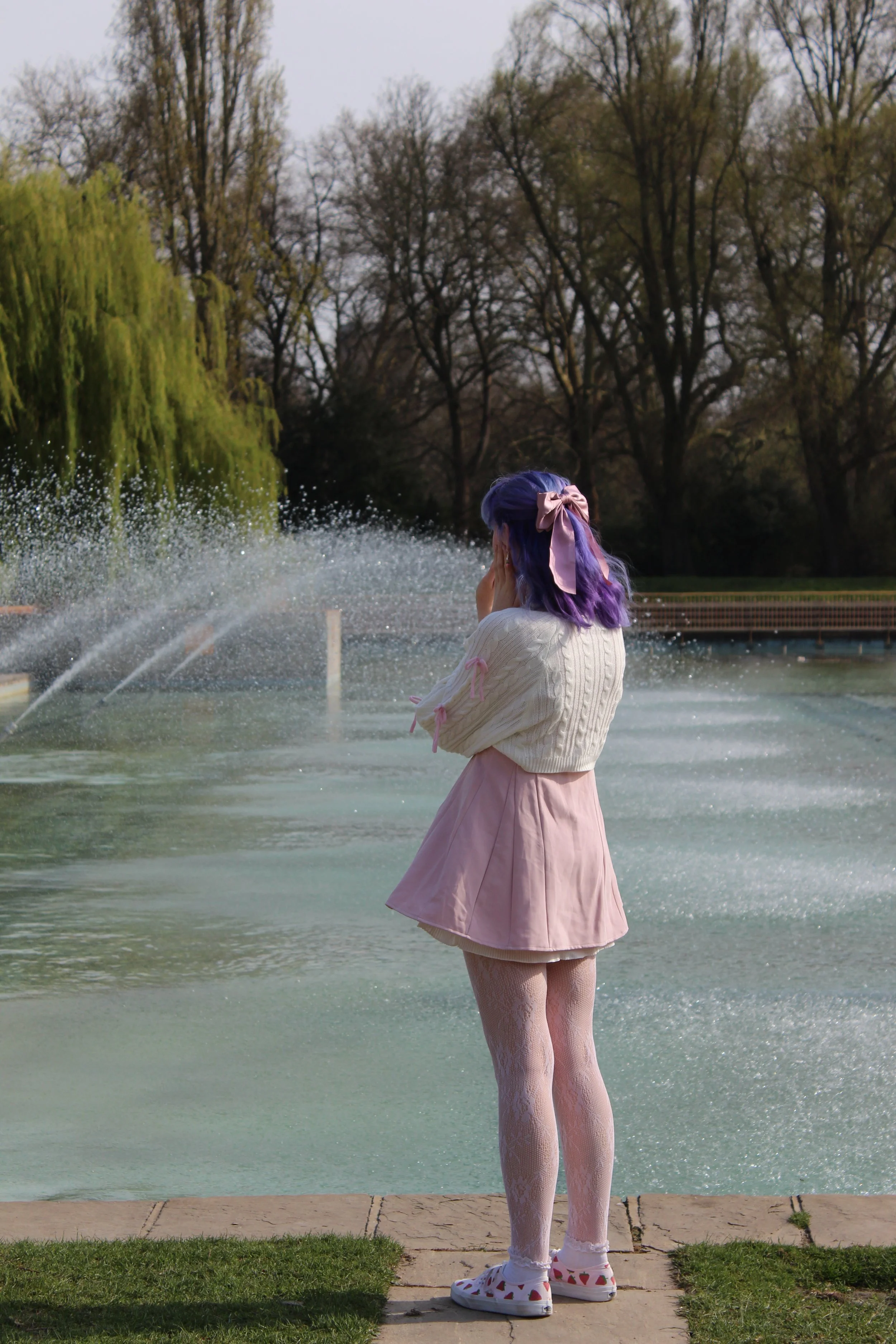 A girl with purple hair and a pink bow stands near water fountain in a park, with trees in the background.
