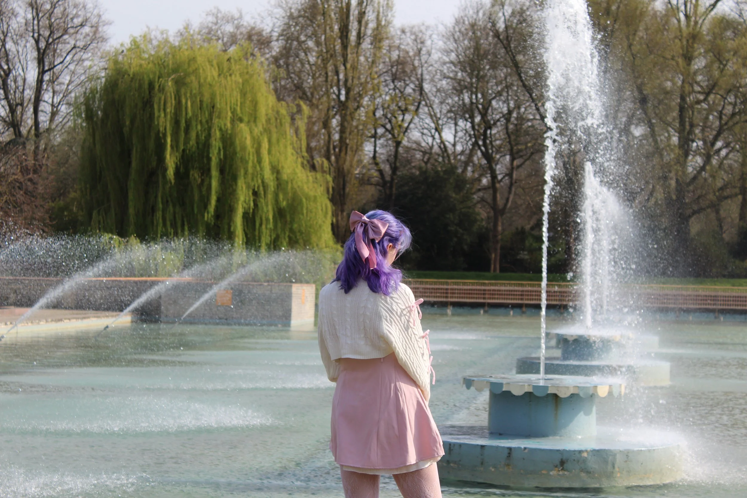 A young girl with long purple hair, tied with a large pink bow, stands near a fountain in a park, surrounded by trees in early spring.