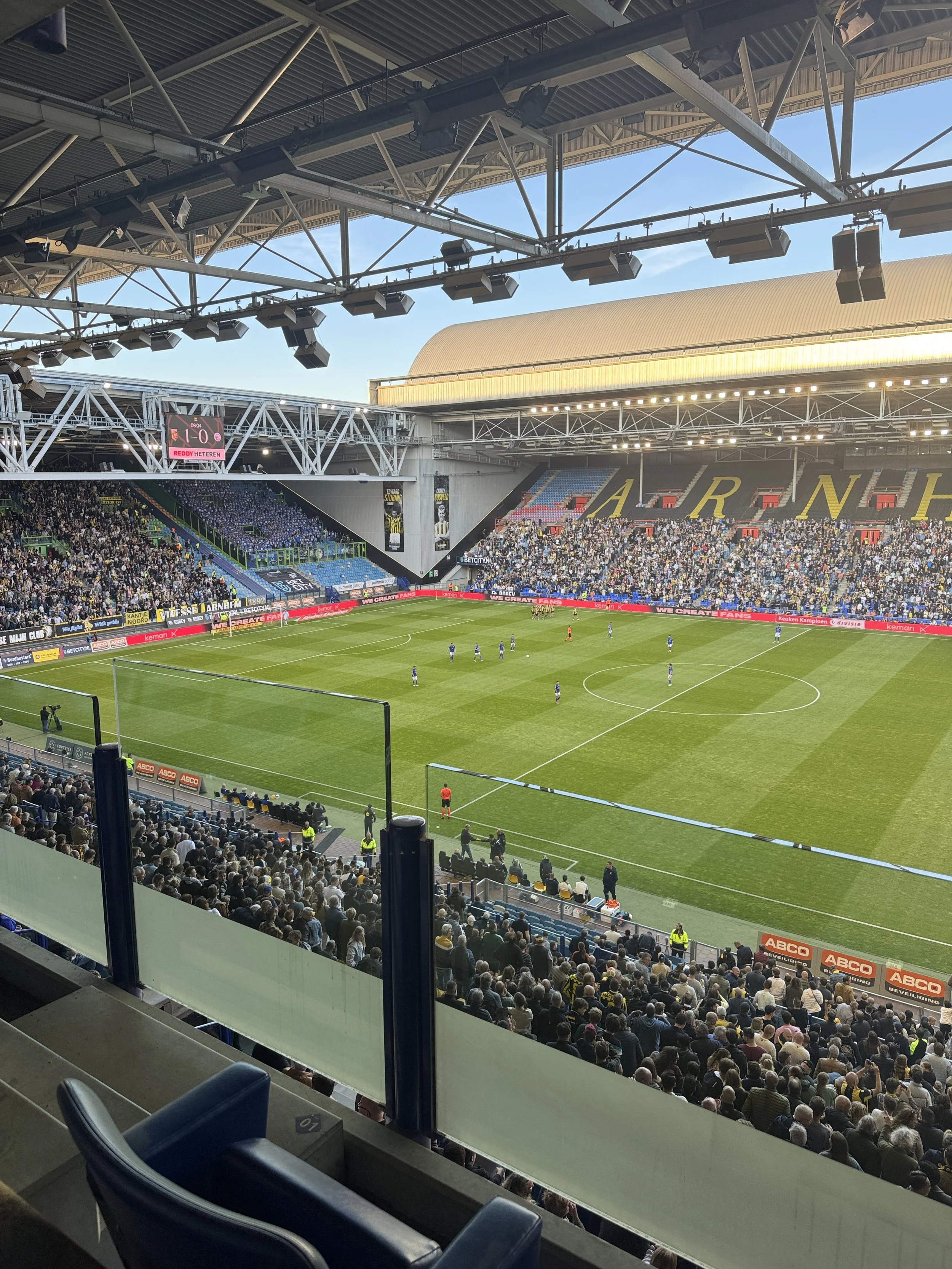 A soccer stadium filled with spectators, with players on the field and bright sunlight illuminating the scene.