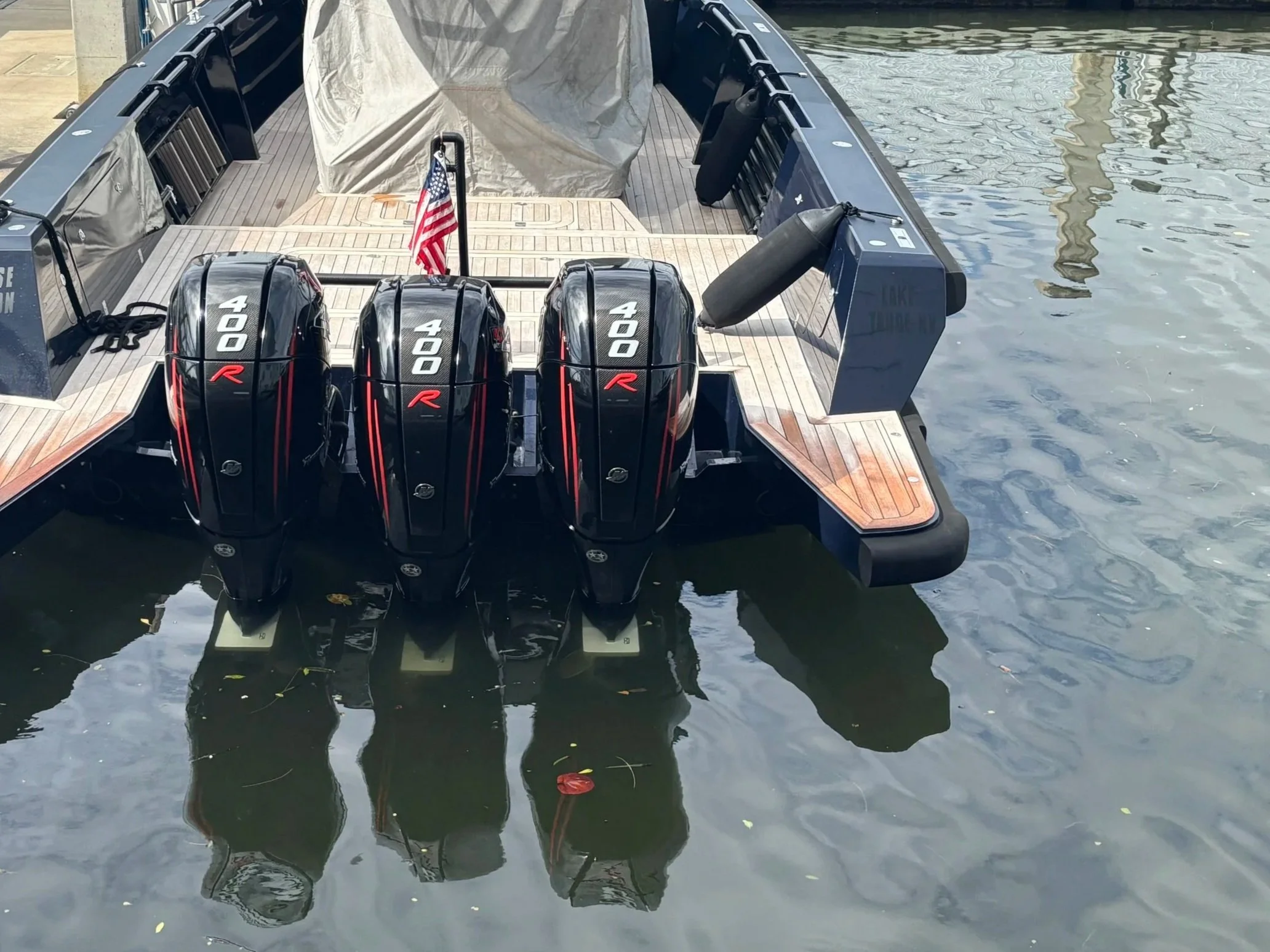 A boat docked at a marina with three black outboard motors labeled '400 R' attached to the stern. The boat has a wooden deck, an American flag, and is floating on water with a reflection.