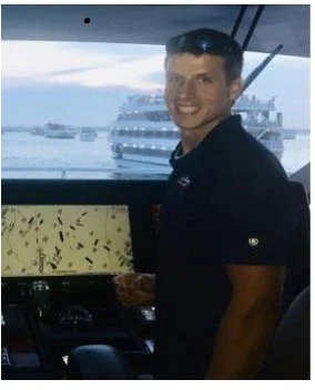 A handsome young man standing on a boat, smiling, with a cruise ship and water visible in the background.