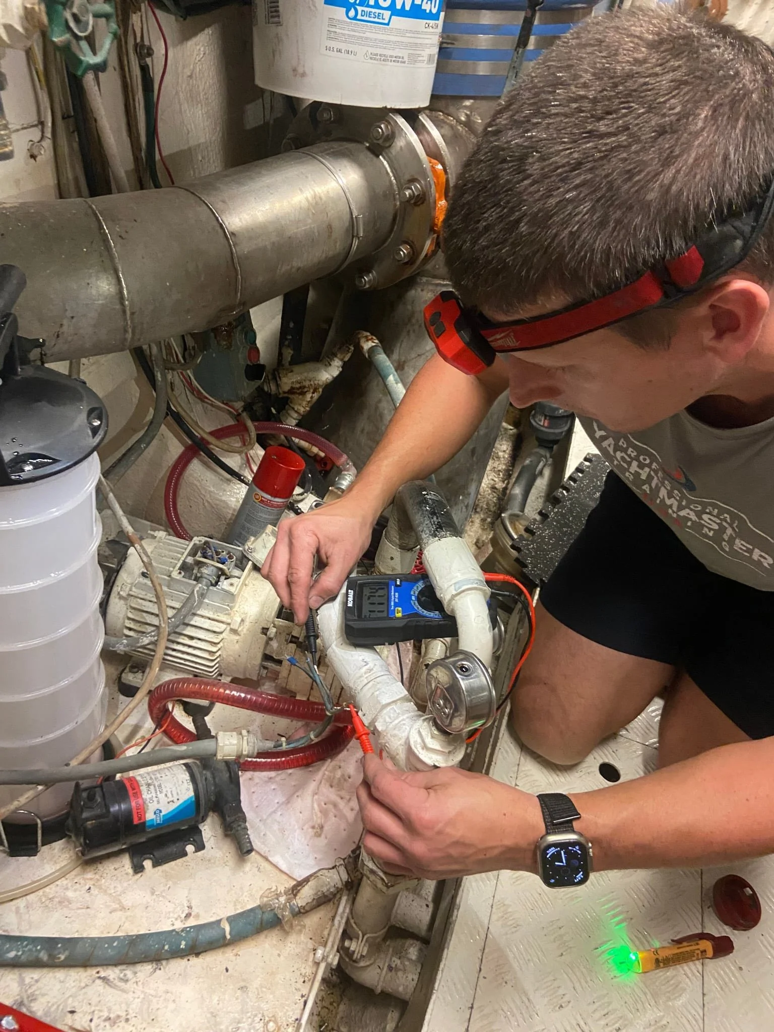 A technician wearing safety glasses  plumbing and equipment involving pipes and a motor, using a digital multimeter, in a mechanical or utility room.