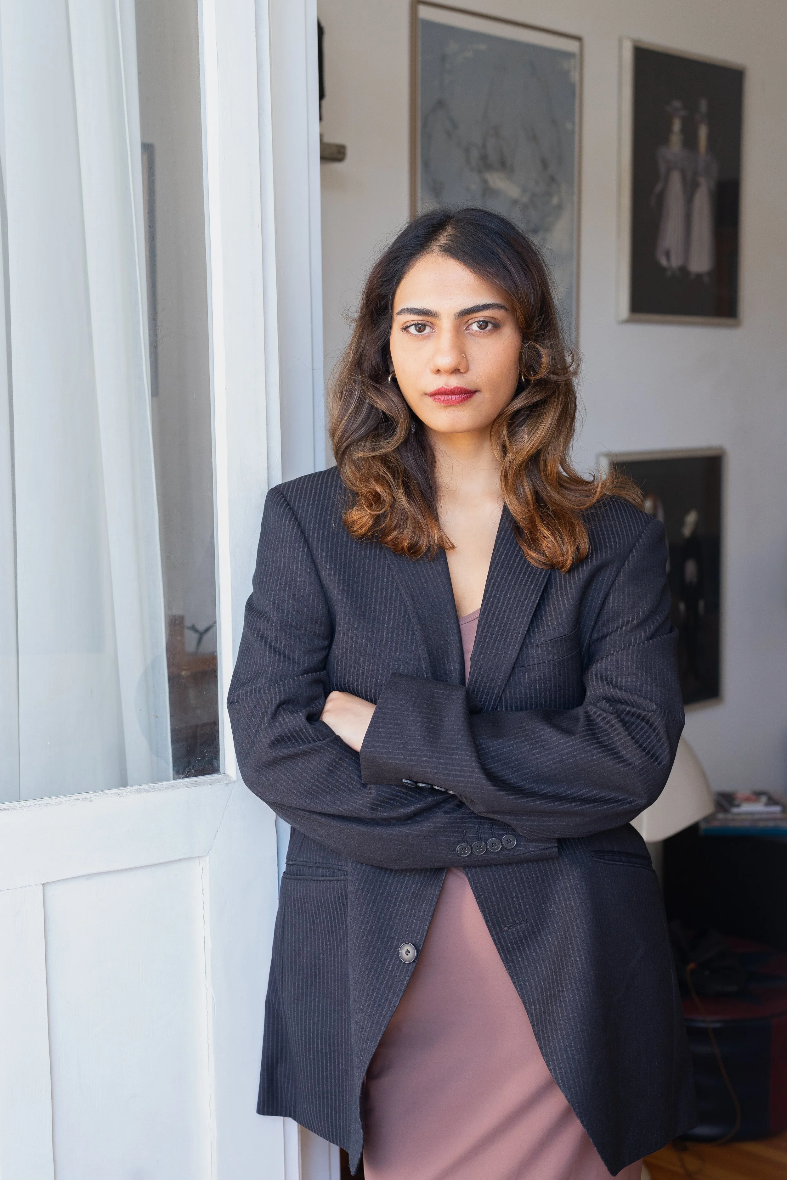 A POC woman psychologist with long hair, wearing a dark blazer and a brown dress, standing with arms crossed by a white doorway, with framed artwork in the background.