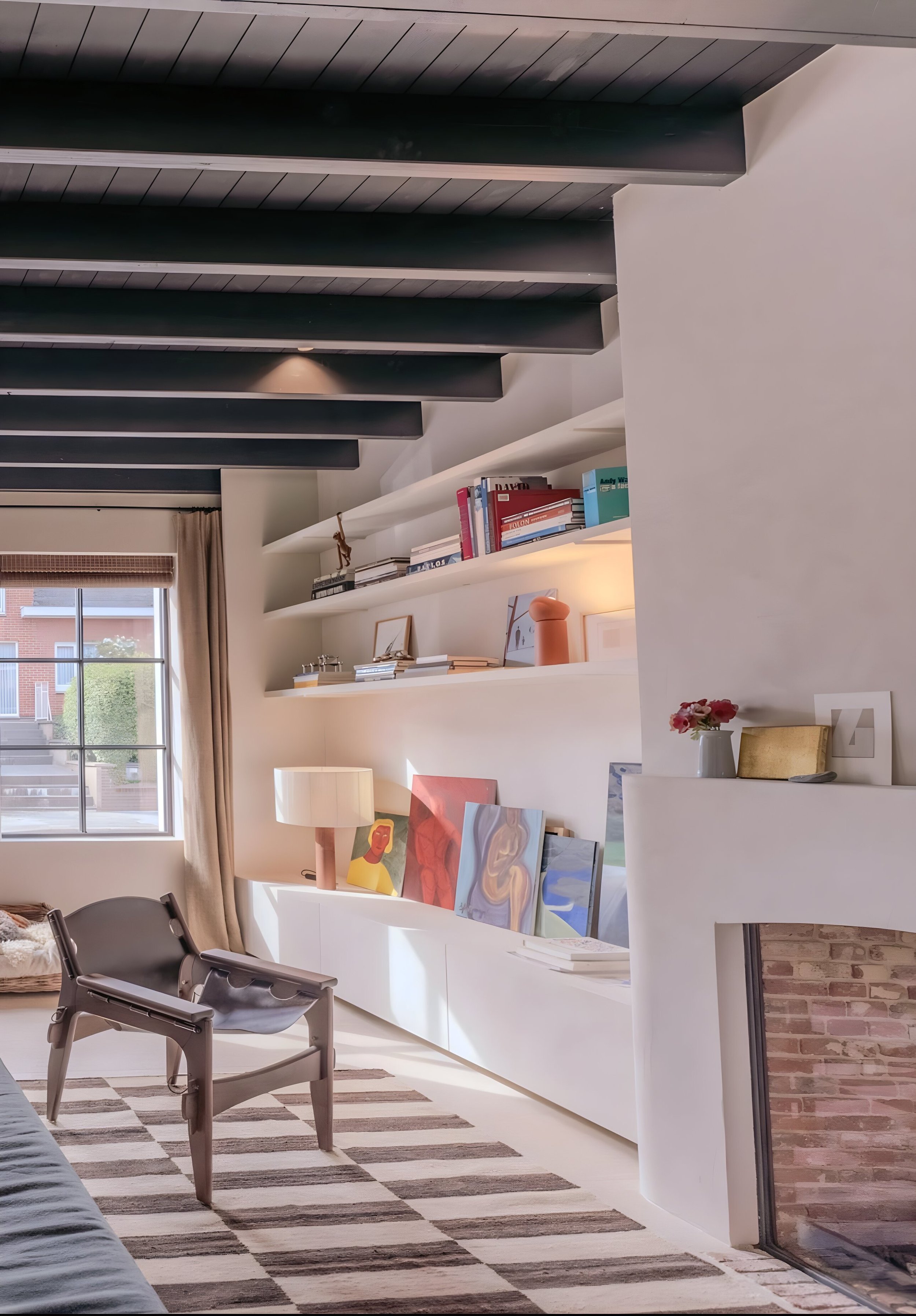 Living room with black ceiling beams, a window with beige curtains, open white shelves with books and art, a lamp with a pink base, and paintings leaning against the wall near a fireplace.