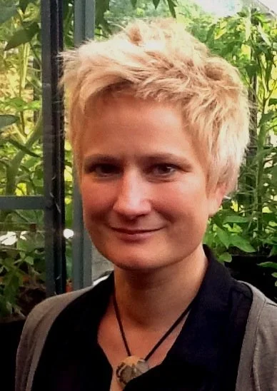 A woman with short, blonde hair, wearing a black shirt and a necklace, standing outdoors surrounded by green plants.