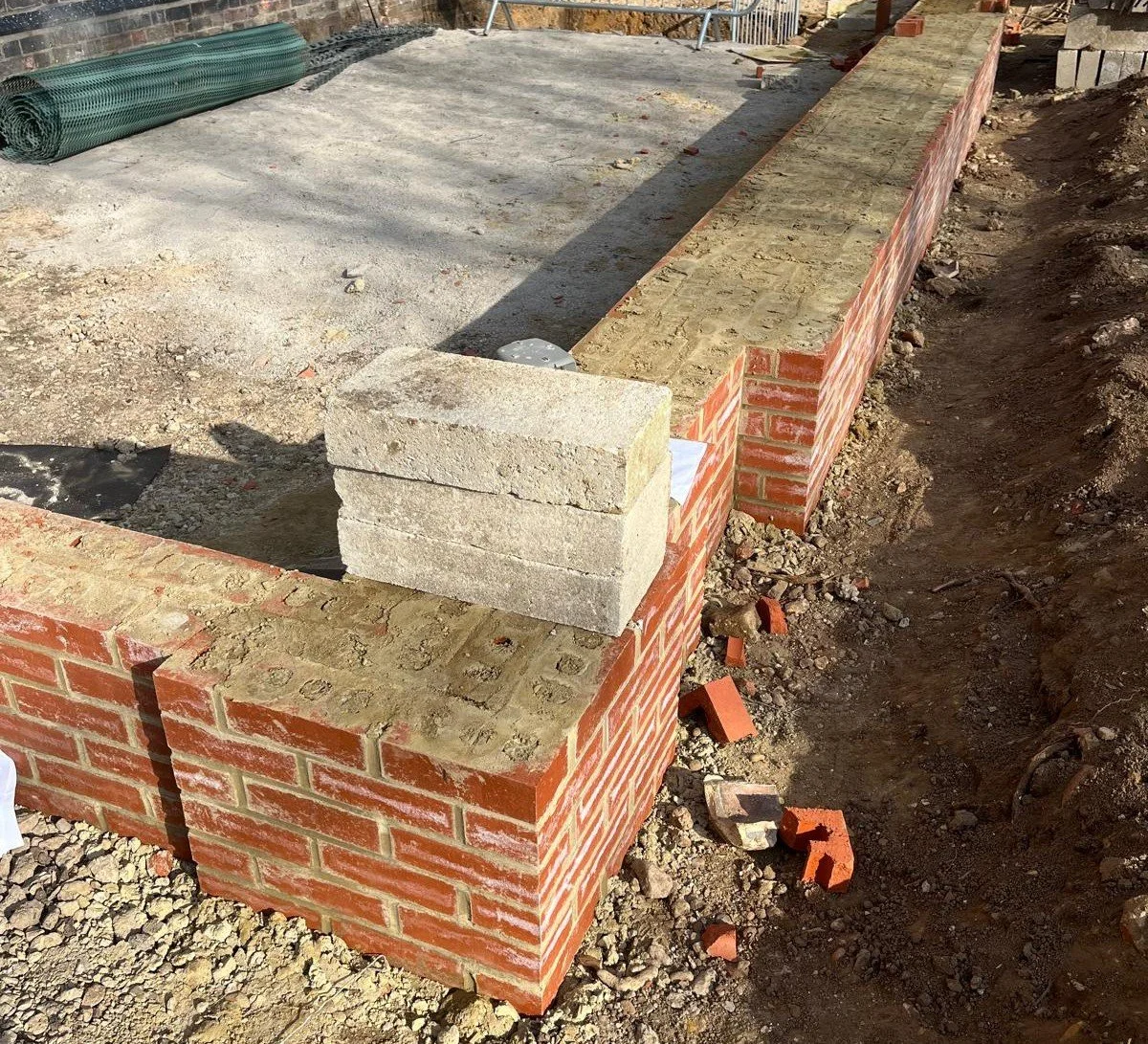 Construction site showing a brick wall foundation being built, with bricks and cinder blocks stacked and partially laid, surrounded by dirt and construction debris.