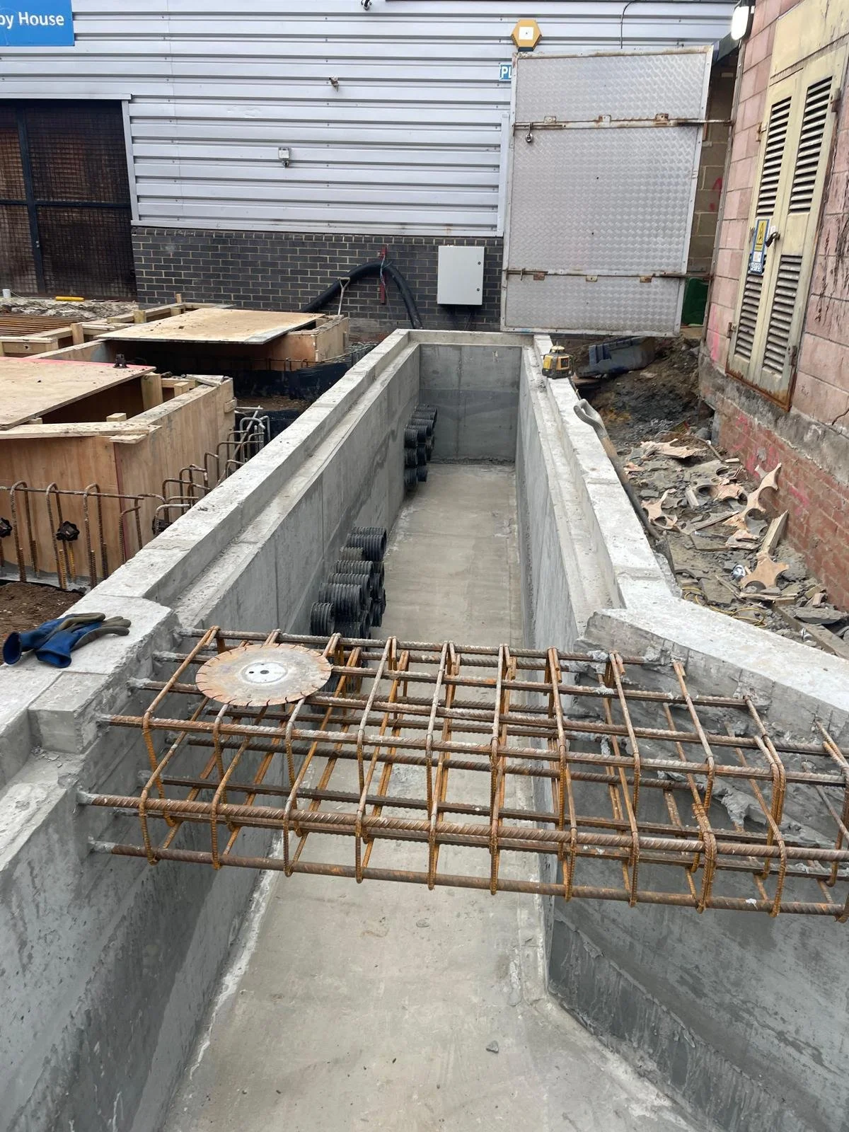 Construction site in South East England showing a partially built concrete pool with rebar and formwork, surrounded by construction materials and equipment.
