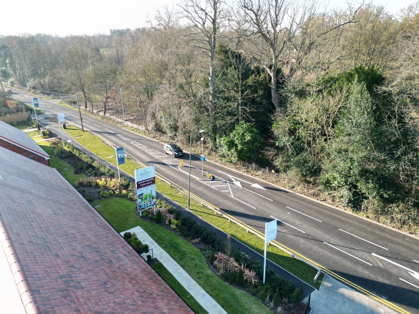 A view from above of a street with parked cars, green landscaped areas, and a surrounding wooded area with leafless trees.