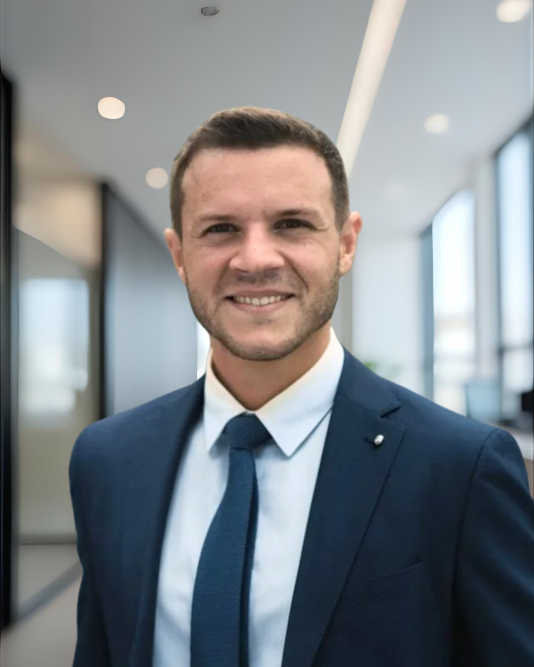 Professional young man in a navy blue suit, white shirt, and navy tie, smiling in a modern office with large windows and bright lighting.